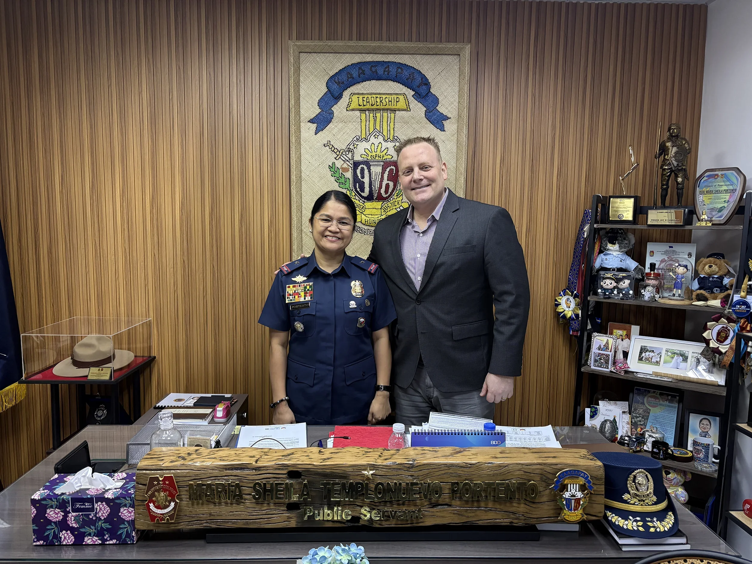 A woman in a police uniform standing next to a man in a suit, both smiling in an office with wooden wall paneling, a police emblem suggests law enforcement connection.