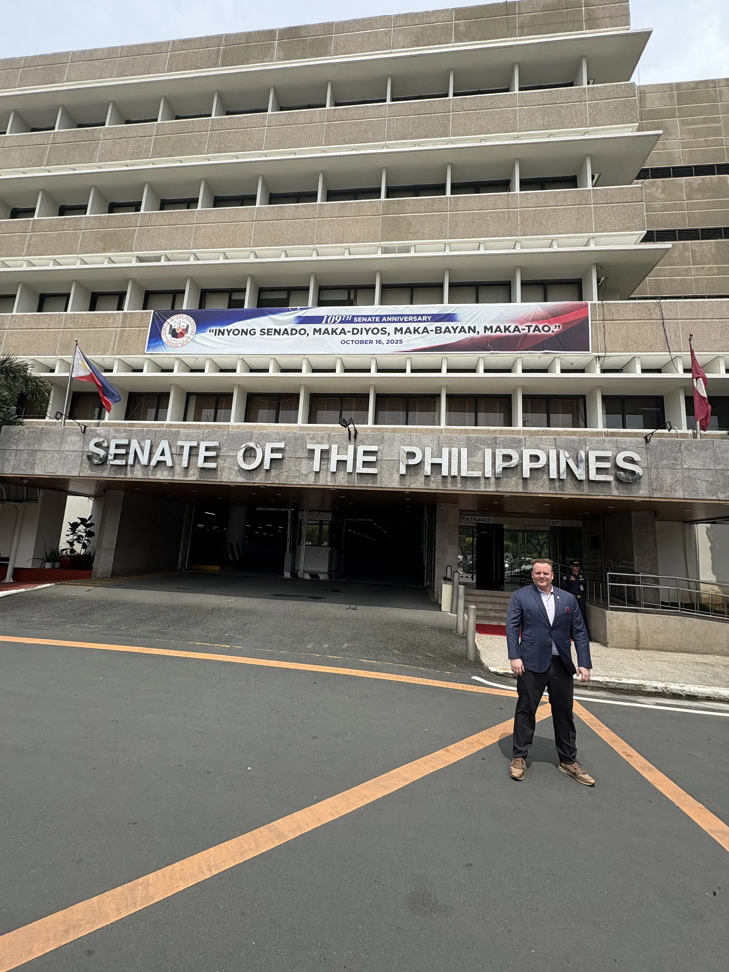 A man in a suit standing outside the entrance of the Senate of the Philippines building, with a man in uniform behind him. The building has a large sign reading "Senate of the Philippines" and a banner celebrating the 109th anniversary of the Senate.