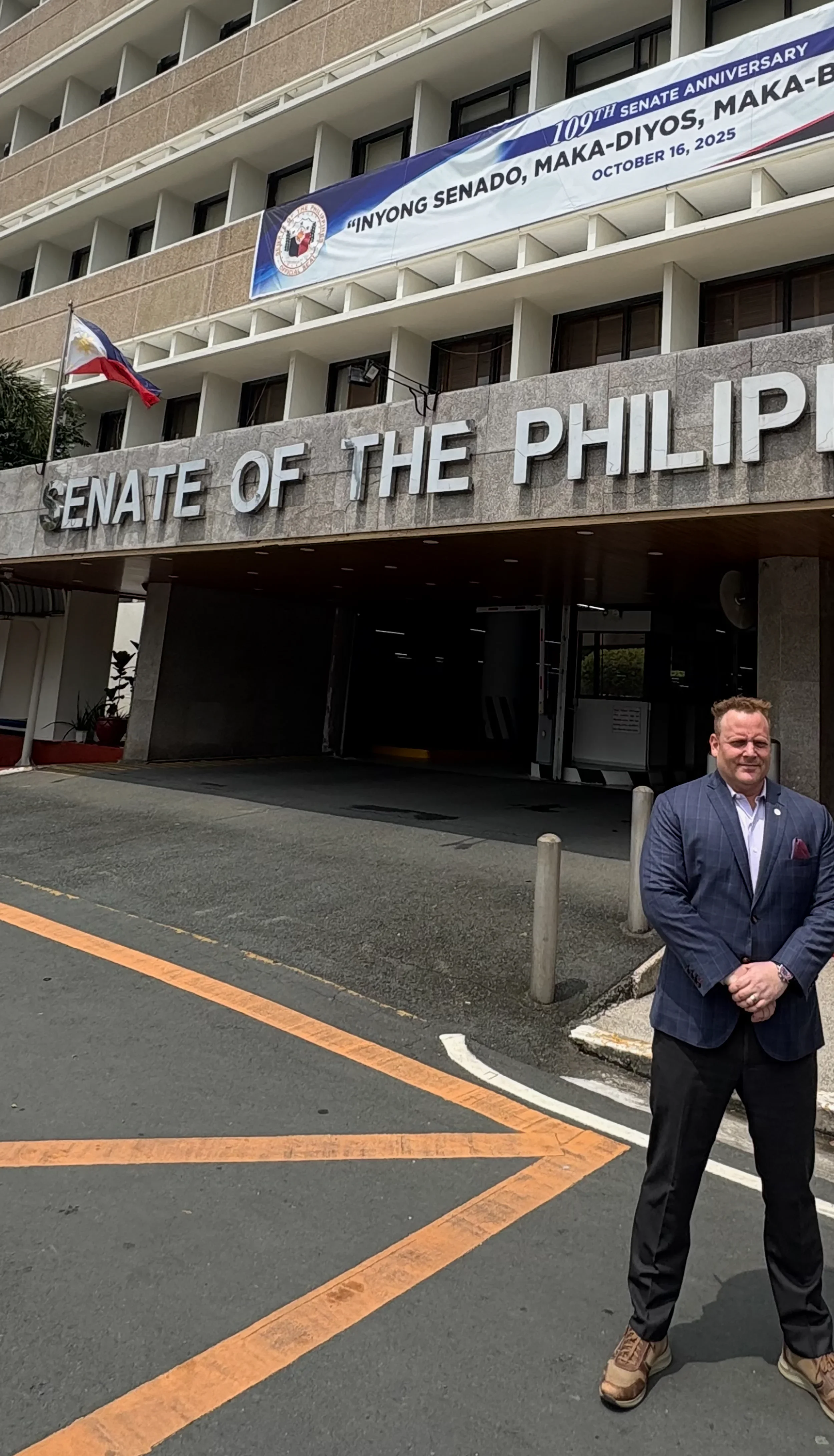 Man standing in front of the Senate of the Philippines building, with a banner above celebrating the 109th Senate anniversary and the Philippine flag to the left.