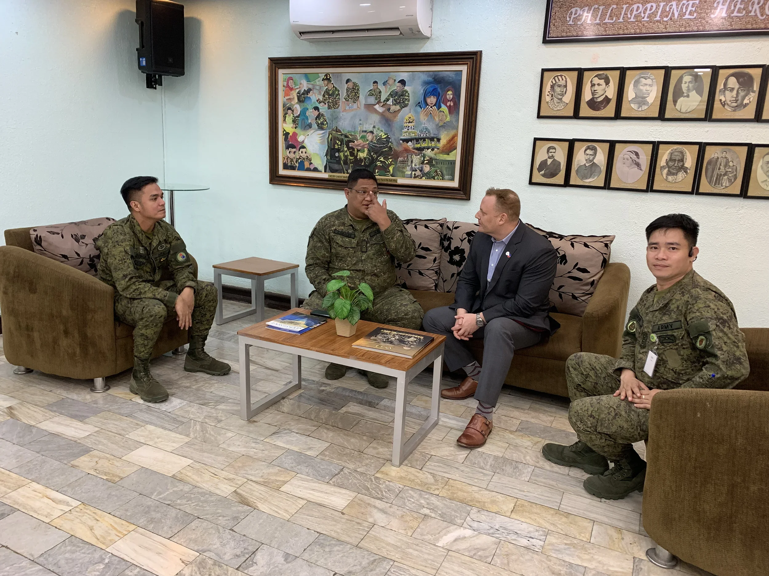 Four men, two in military uniforms and two in formal civilian attire, sitting on sofas and engaging in a discussion in a room with framed pictures and a colorful mural on the wall.