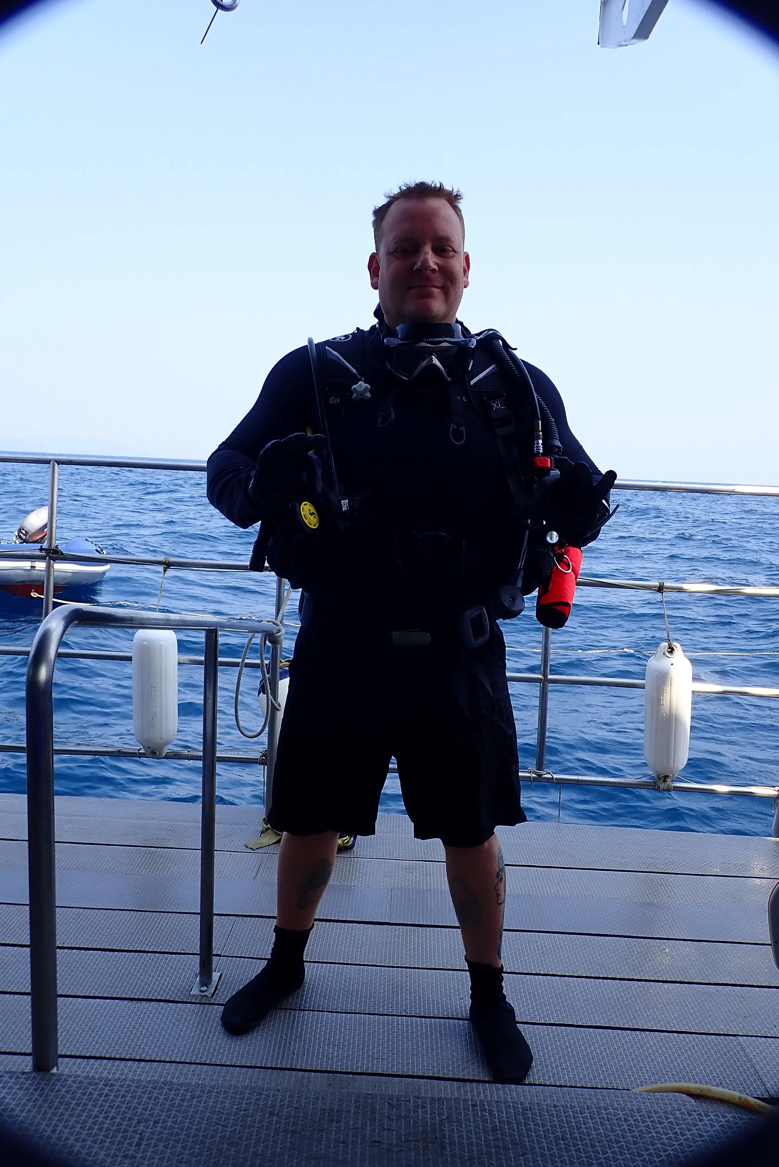 A man in scuba gear standing on the deck of a boat over the ocean, smiling at the camera.