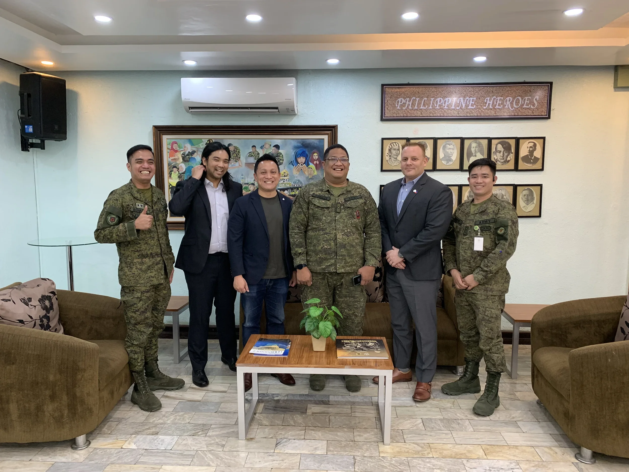 A group of six men, including four in military uniform and two in civilian suits, standing and smiling in a room with framed portraits and a wall sign that says 'Philippine Heroes'.