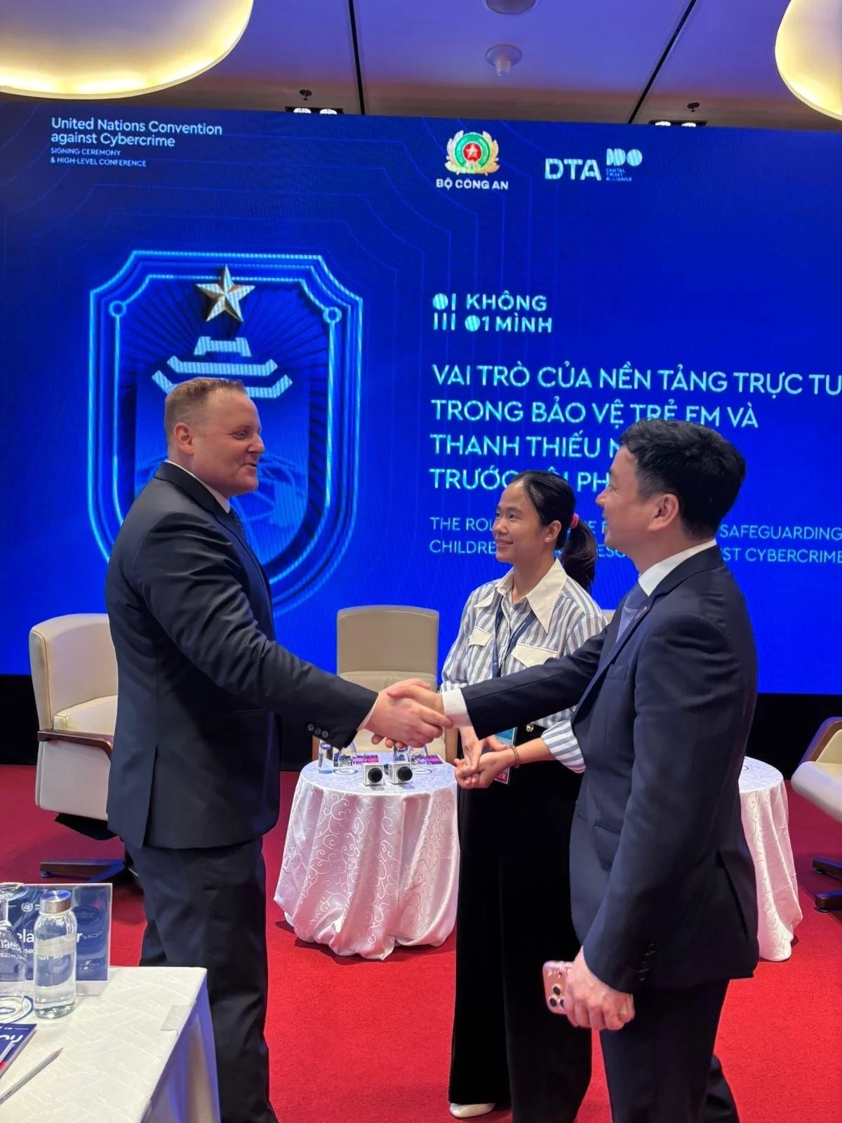 Two men shaking hands in a formal setting during a conference, with a woman standing between them. The background features a large screen displaying text about the United Nations Convention against Cybercrime, law enforcement logos, and conference de
