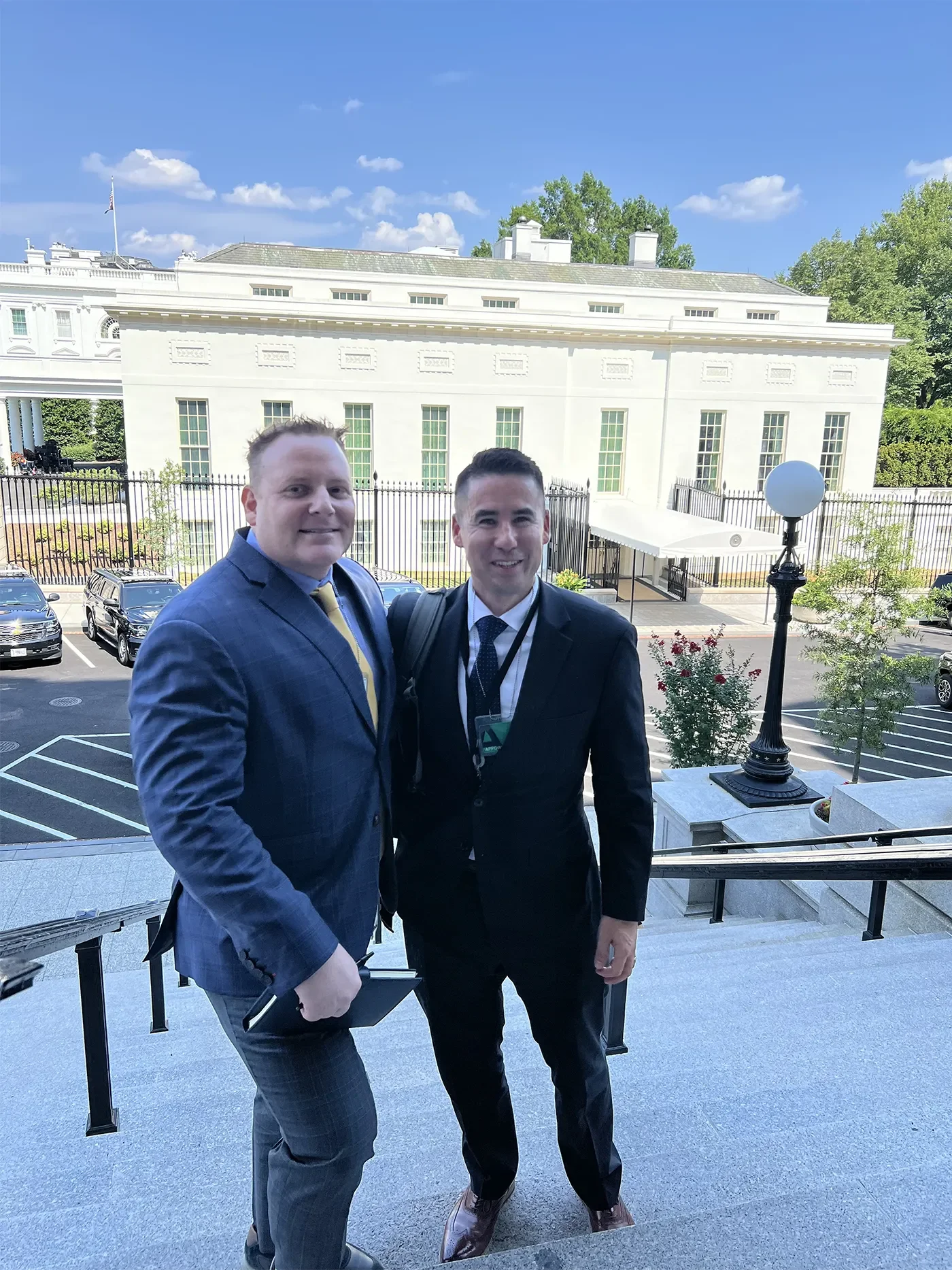 Two men standing on stairs outside of the White House, dressed in business attire, smiling at the camera. One man is wearing a blue checkered suit with a yellow tie, and the other is in a black suit with a blue tie, with an ID badge hanging around hi