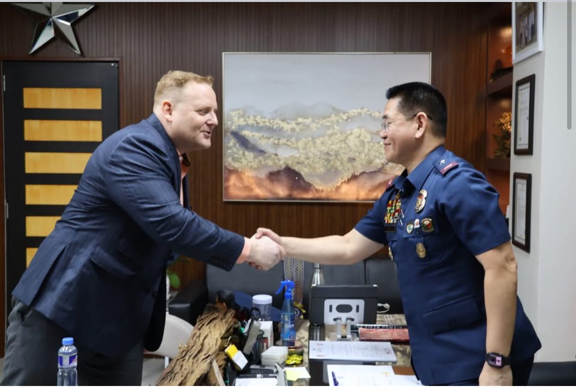 Two men shaking hands in an office; one man in business attire, the other in a police uniform with medals, in front of a desk with papers, bottles, and office supplies.