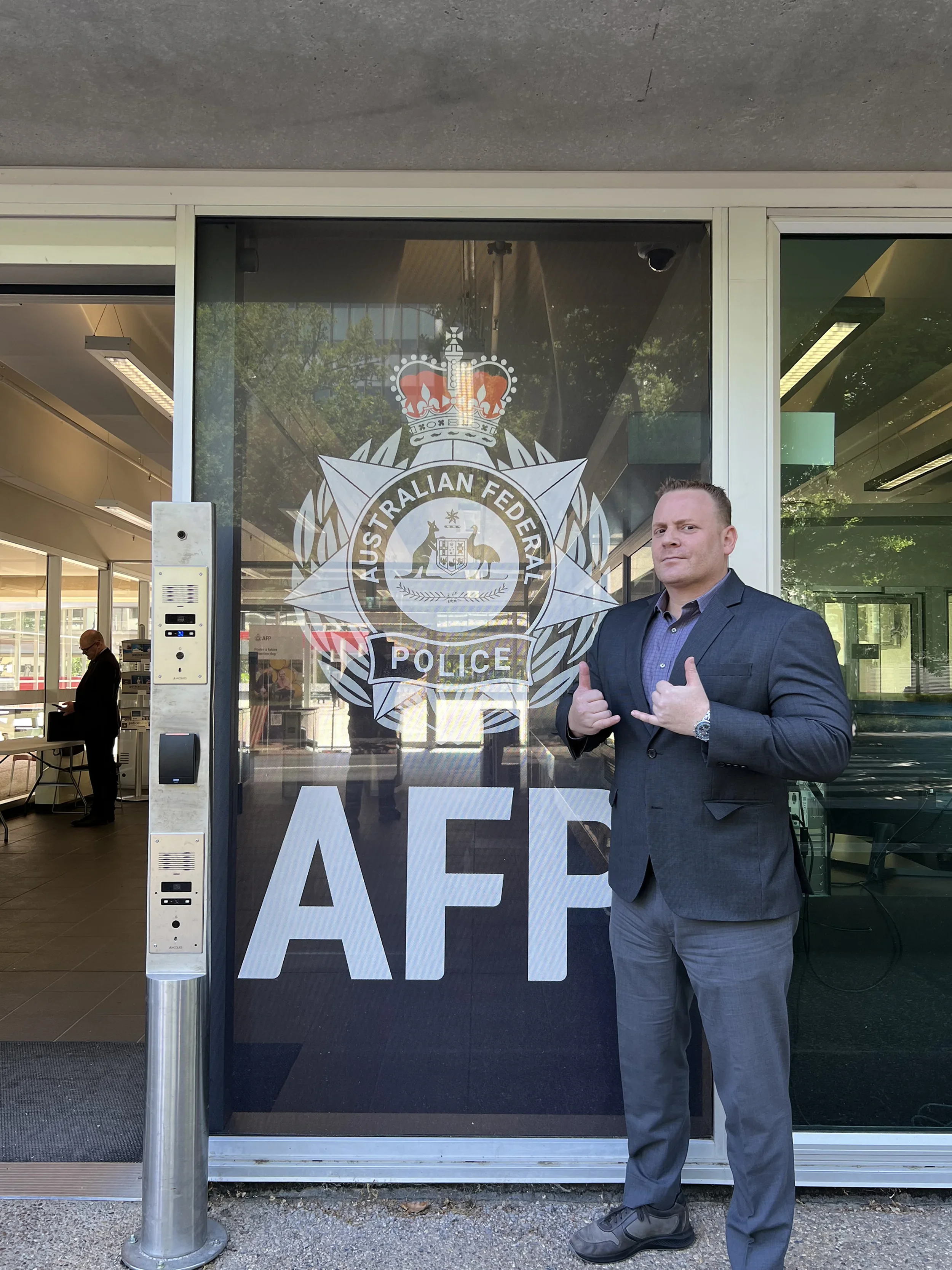 A man in a suit standing outside the Australian Federal Police headquarters, posing with a confident thumbs-up gesture.