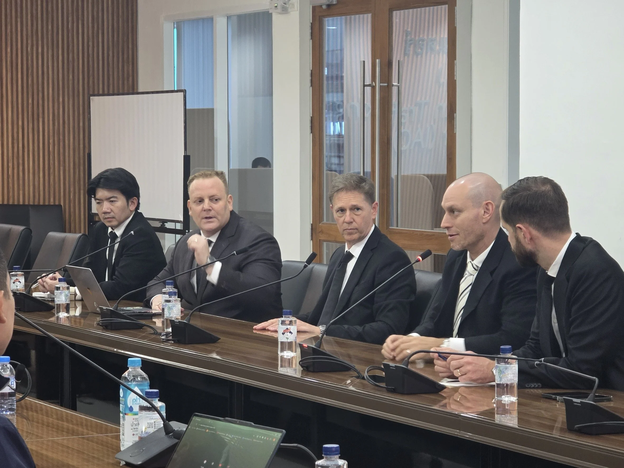 Five men in suits sit at a conference table equipped with microphones, water bottles, and laptops, in a modern meeting room with wooden wall paneling, a whiteboard, and wood-framed glass doors.