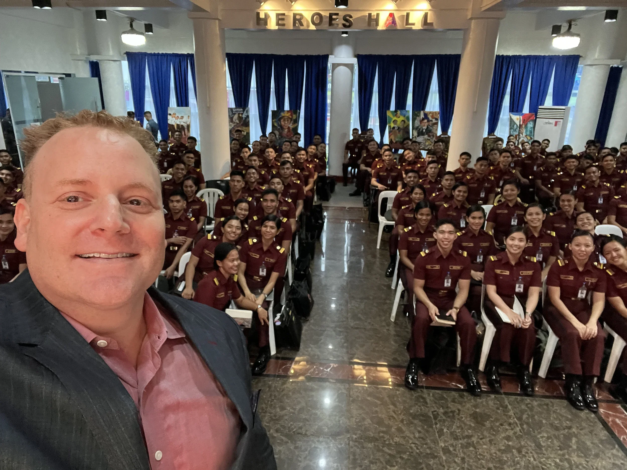 A man taking a selfie in front of a large group of uniformed women seated in an auditorium. The background has blue curtains and signs that say 'Heroes Hall'.