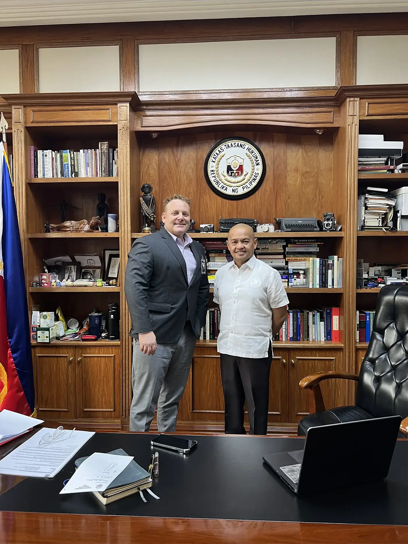 Two men stand in an office in front of a wooden bookshelf with the seal of the Philippines on it. One man is wearing a dark suit and the other is in a white shirt. There are books, photos, and decorations on the shelves, and a desk with documents, a 