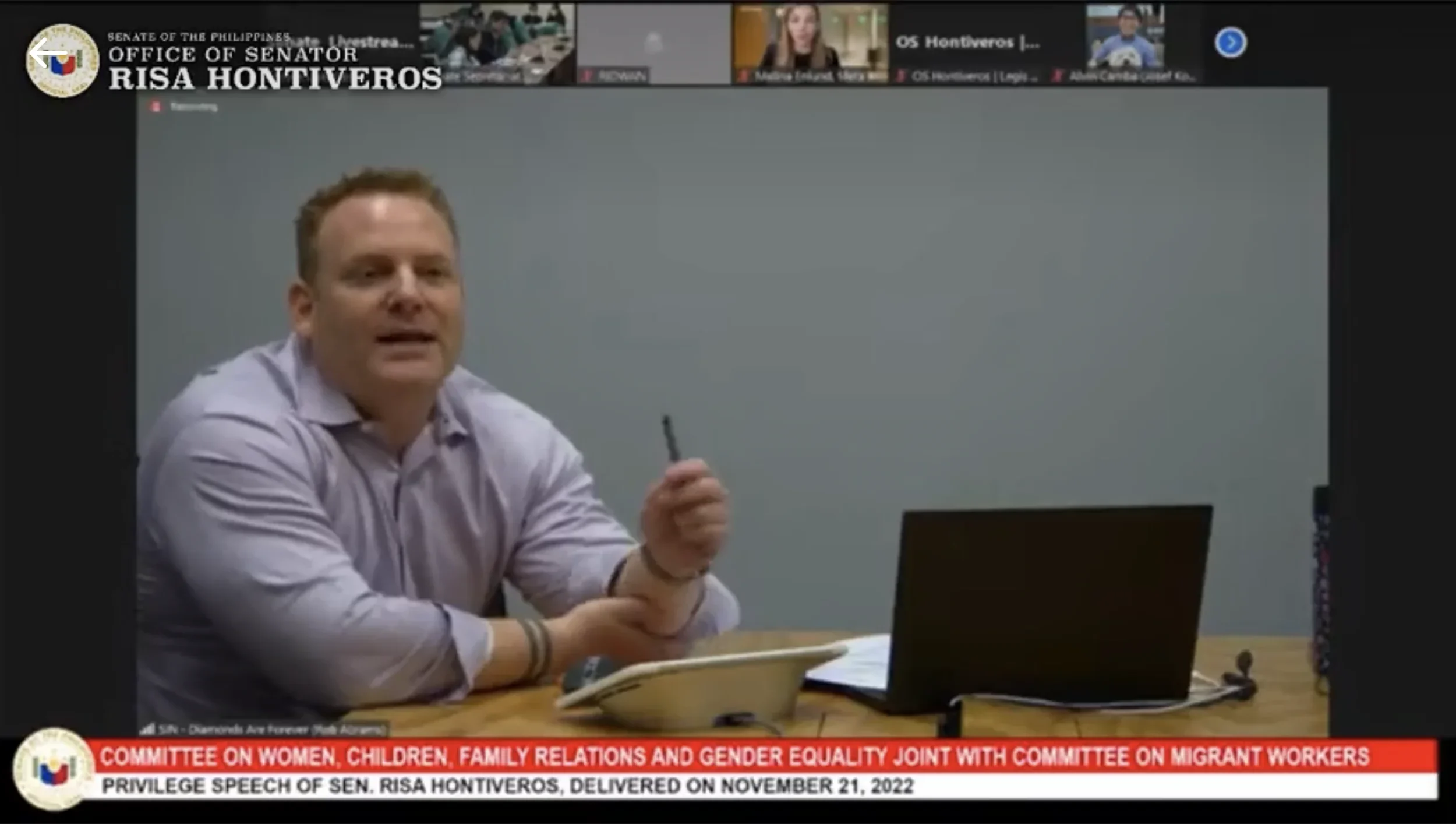 Man speaking during a virtual meeting, with a laptop, phone and papers on the table in front of him, with the text overlay indicating a government committee meeting.