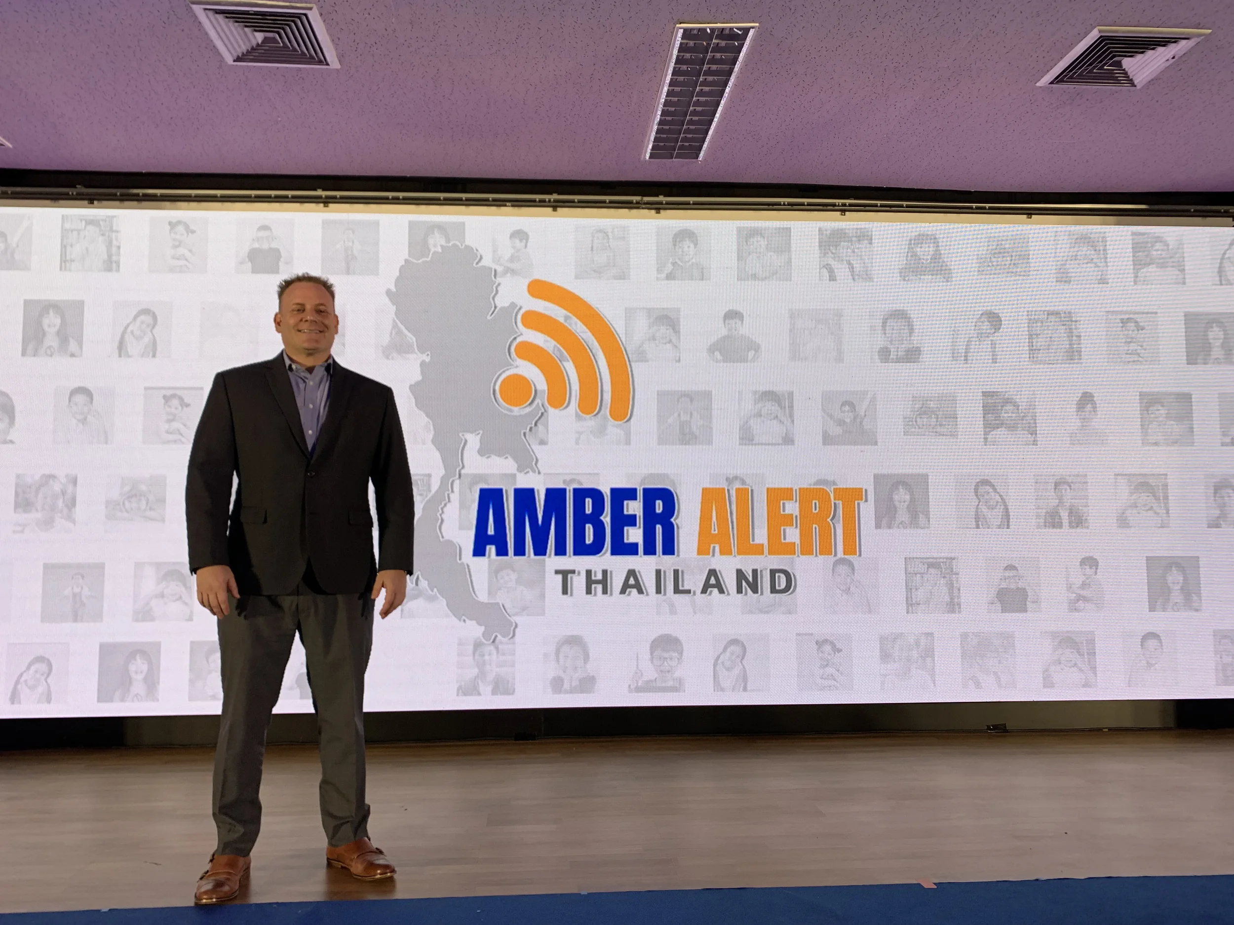 A man in a dark suit and dress shoes standing in front of a large screen displaying the Amber Alert Thailand logo, which includes a map of Thailand with a radio signal icon above it, and a mosaic of black-and-white photographs of children's faces in 