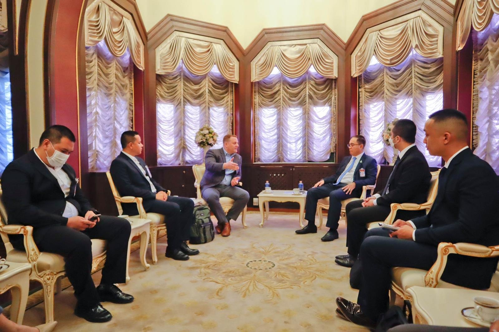 A group of six men in suits seated in a formal room, engaged in discussion; some are wearing face masks and one is using a smartphone.