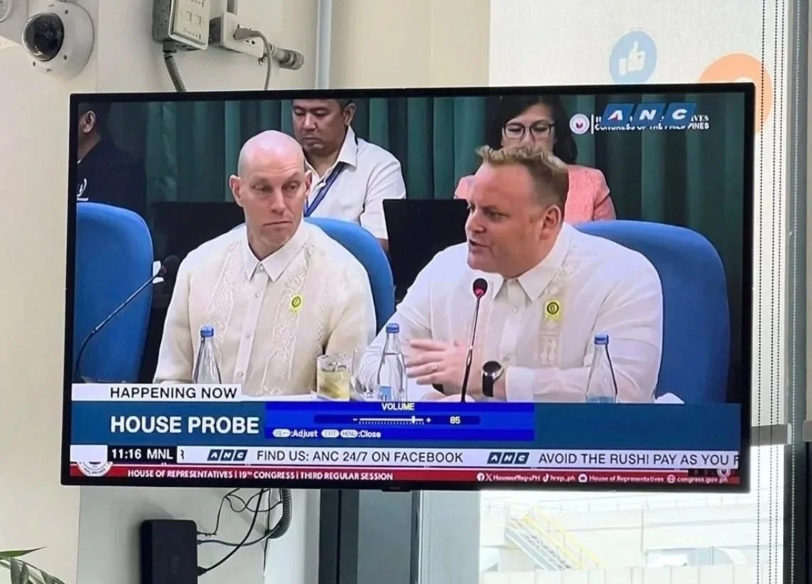 A man taking a selfie in front of a large group of people dressed in matching maroon uniforms in a hall. The hall has blue curtains and various colorful posters on the walls. The background has a sign reading 'HEROES HALL'.