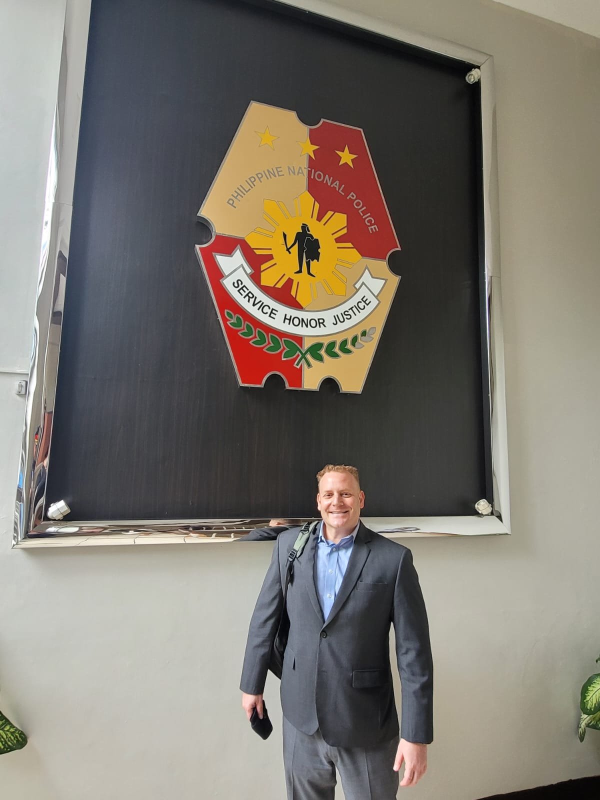 Man in a gray suit standing in front of a large Philippine National Police emblem on a wall.