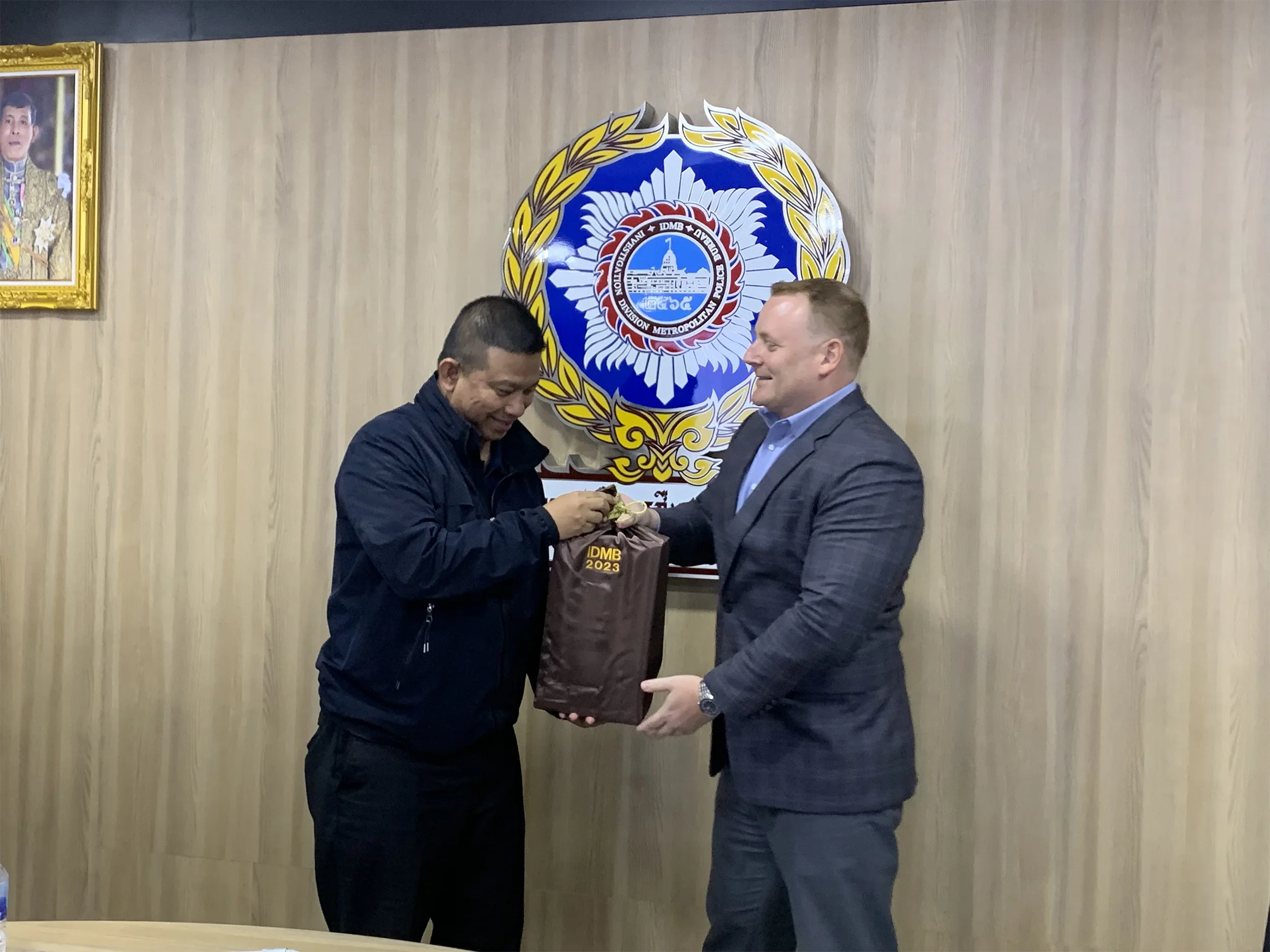 Two men exchanging a gift during a formal meeting in front of a police emblem. One man is wearing a black jacket, the other a gray suit. A portrait hangs on the wall to the left.