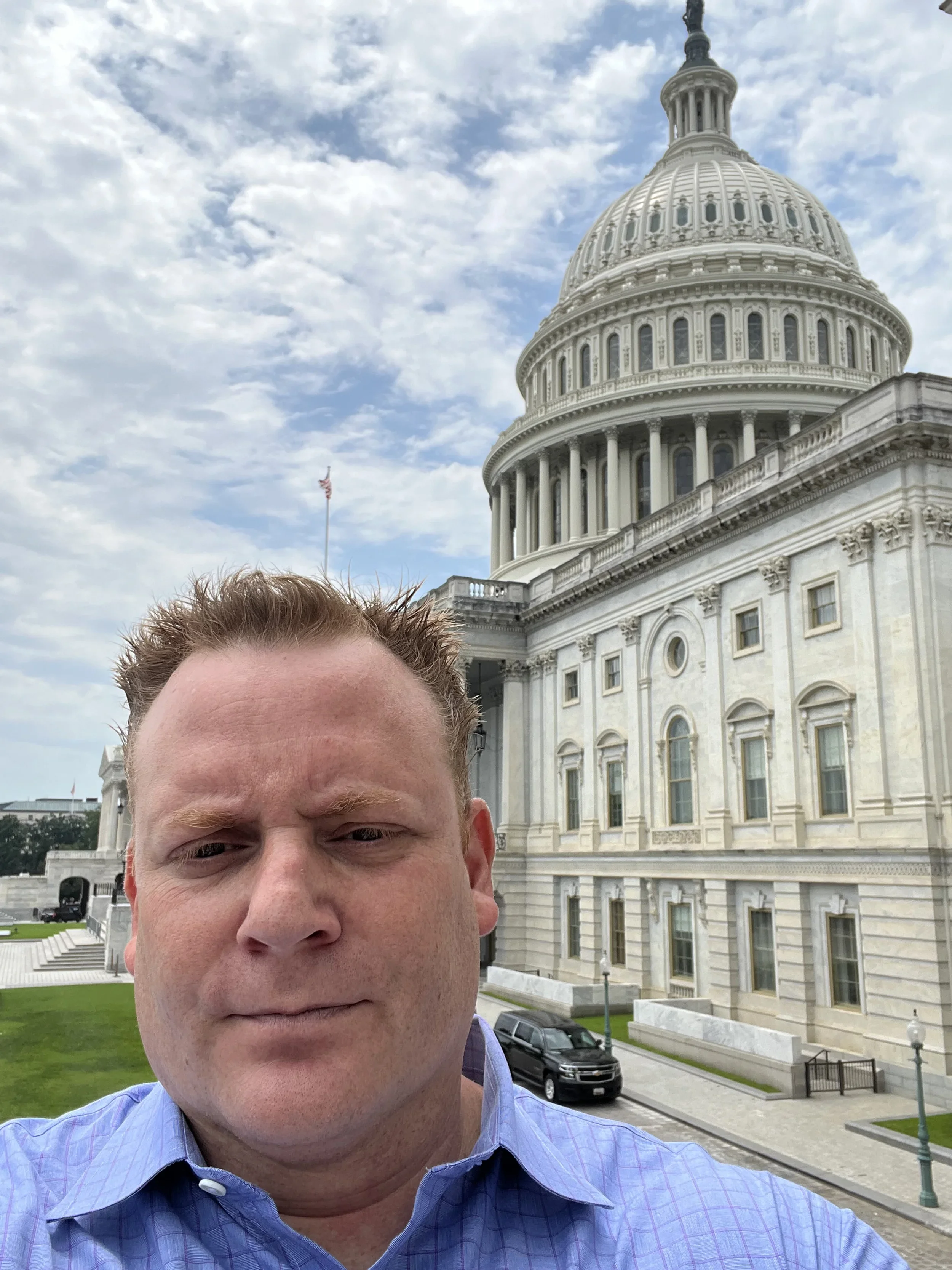 A man taking a selfie in front of the United States Capitol building with a partly cloudy sky in the background.