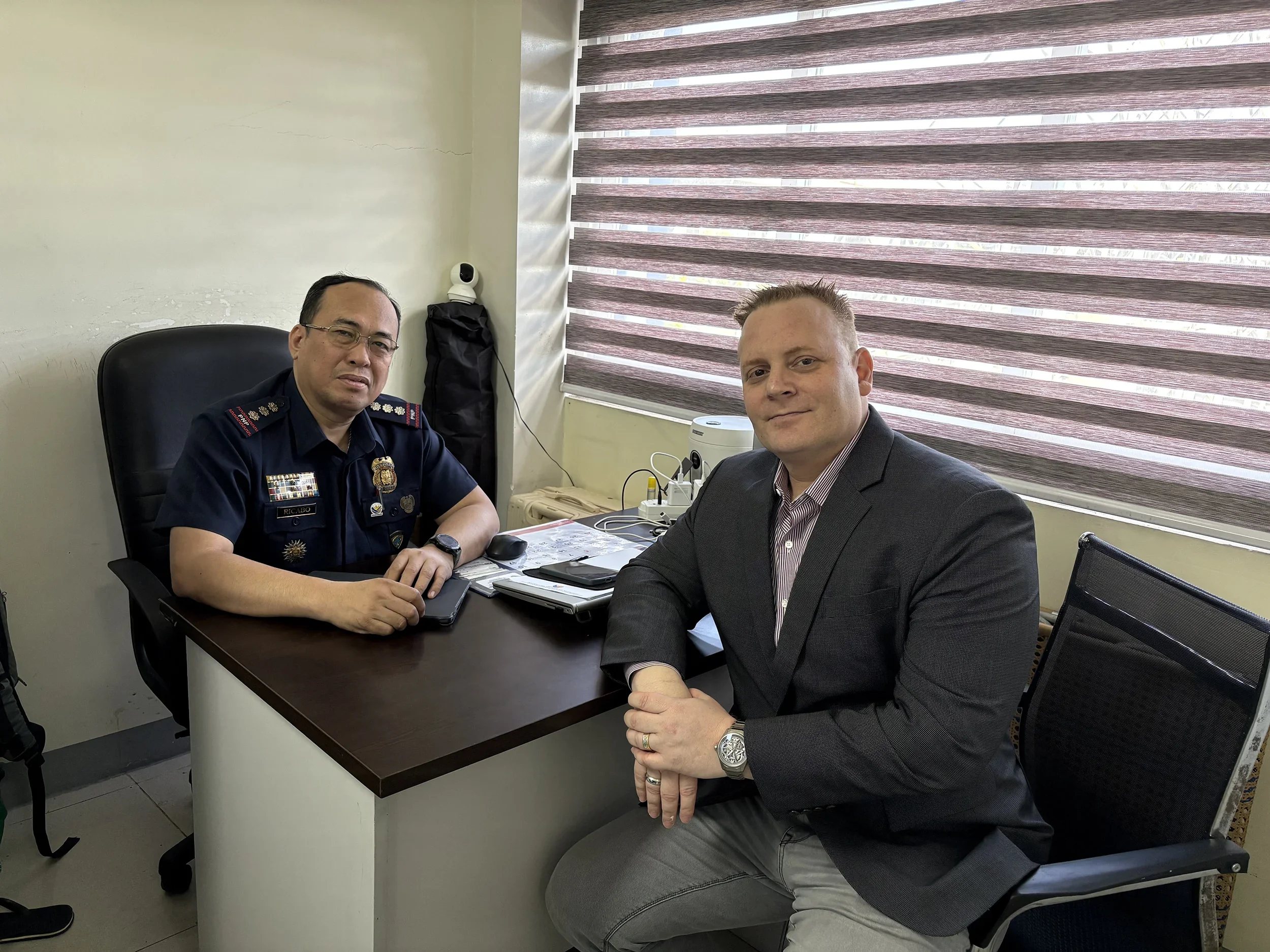 Two men sitting in an office, one in a police uniform and the other in a business suit, with a desk, papers, and blinds in the background.