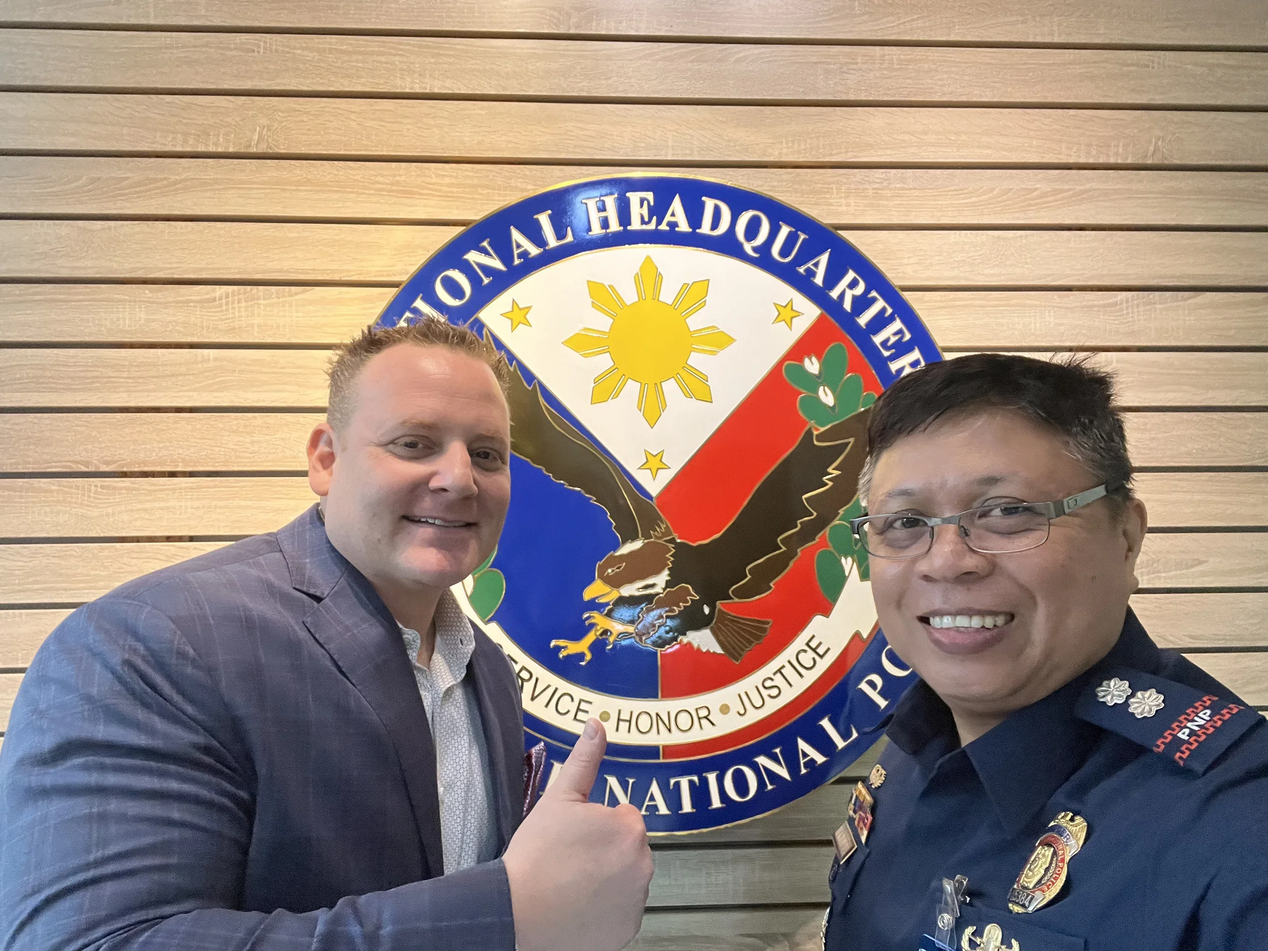 Two men smiling in front of a logo for the National Headquarters. The man on the left is wearing a purple suit jacket and pointing to the logo. The man on the right is wearing a police uniform with various badges and is also smiling.