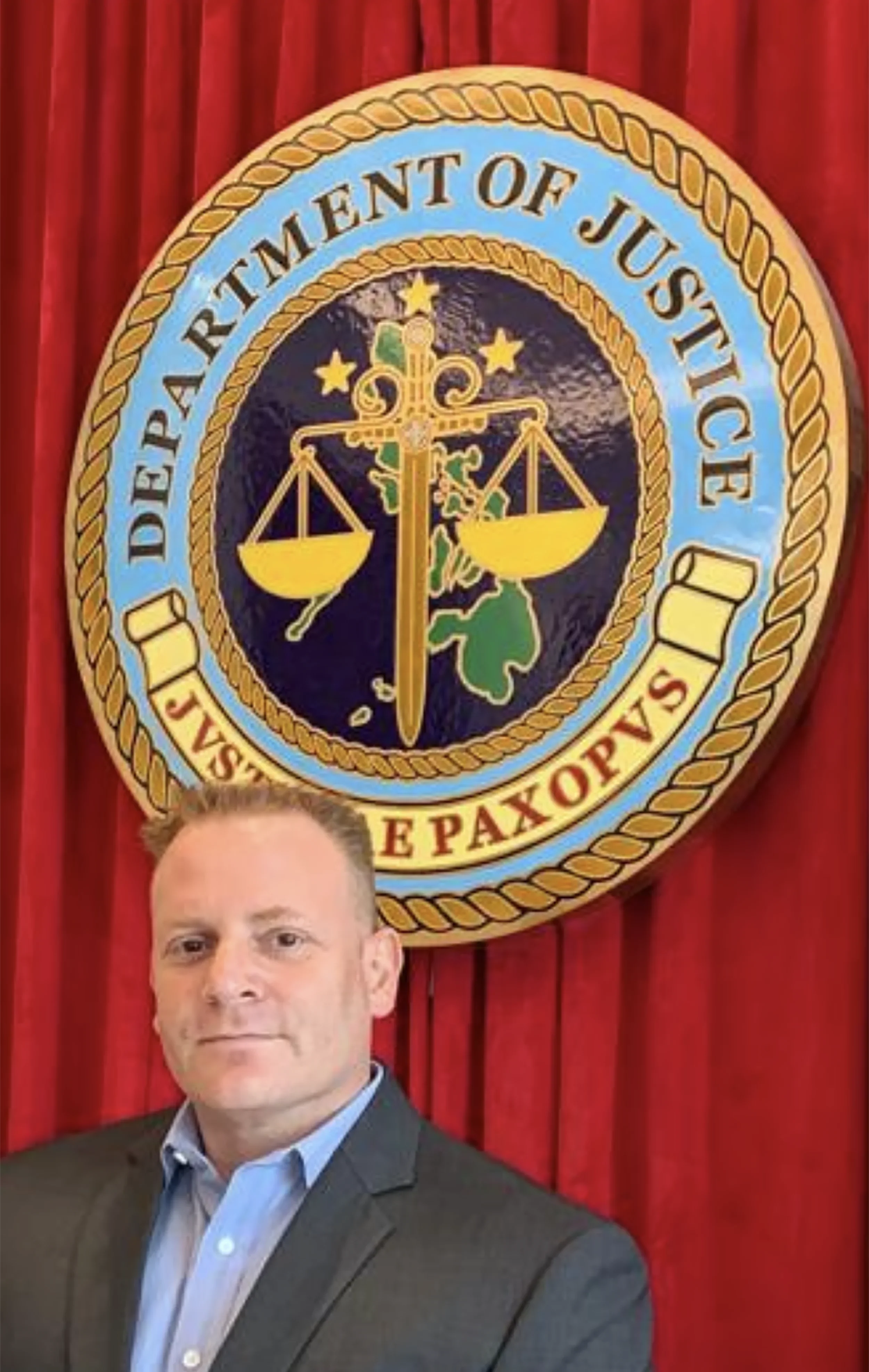 Man in a suit standing in front of the Department of Justice emblem with red curtains backdrop.