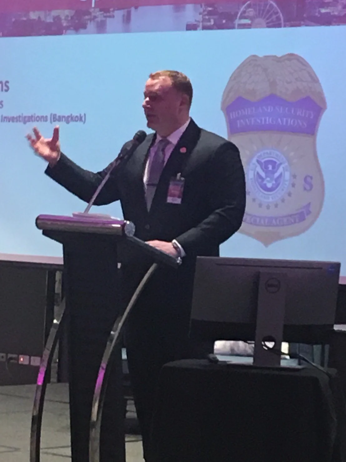 A man in a suit and tie giving a presentation at a podium with a microphone. Behind him is a screen displaying a badge with the words 'Homeland Security Investigations' and a logo.