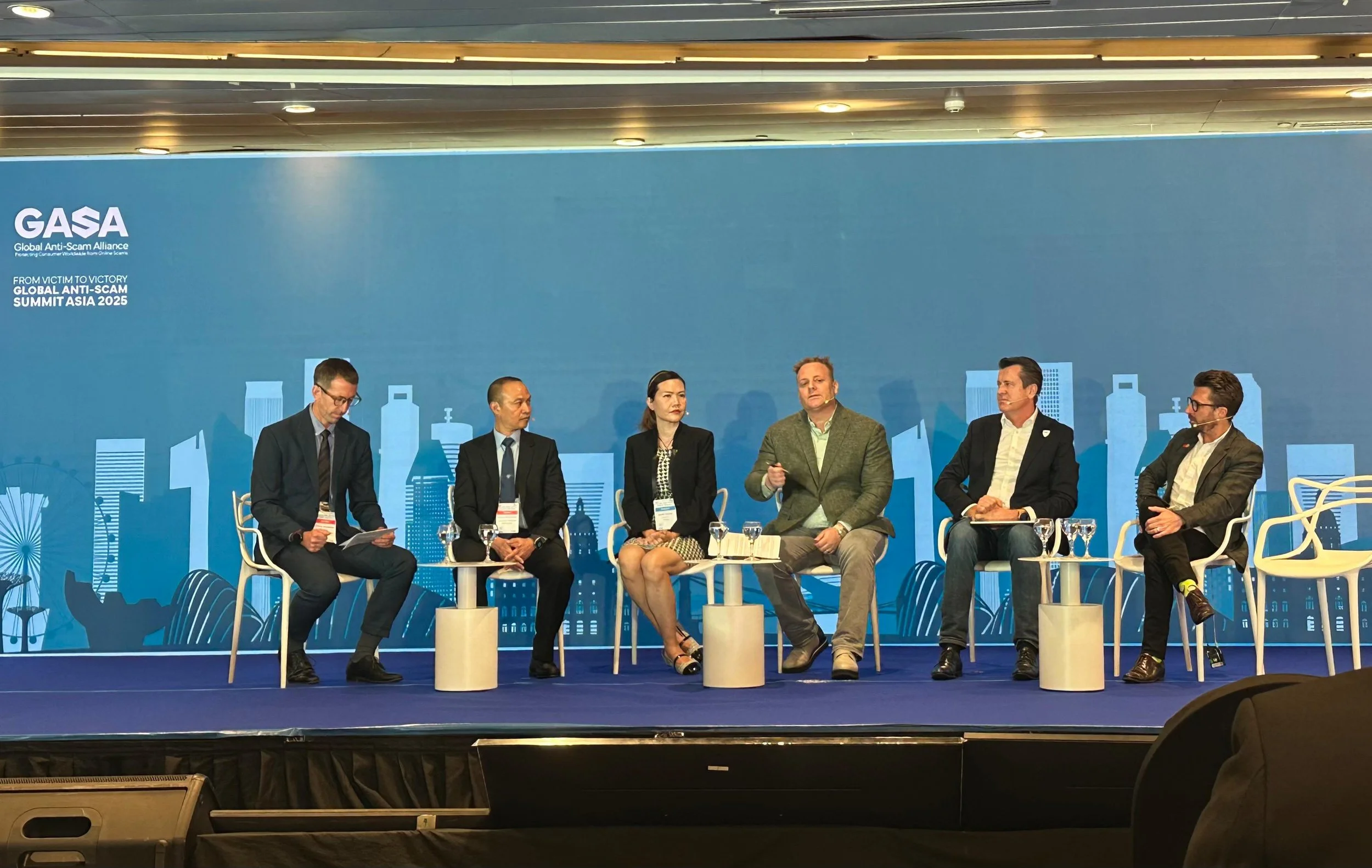 Six people sitting on a stage in a panel discussion at the Global Anti-Scam Summit Asia 2025, with a blue cityscape backdrop, six glasses of water, and papers on their laps.