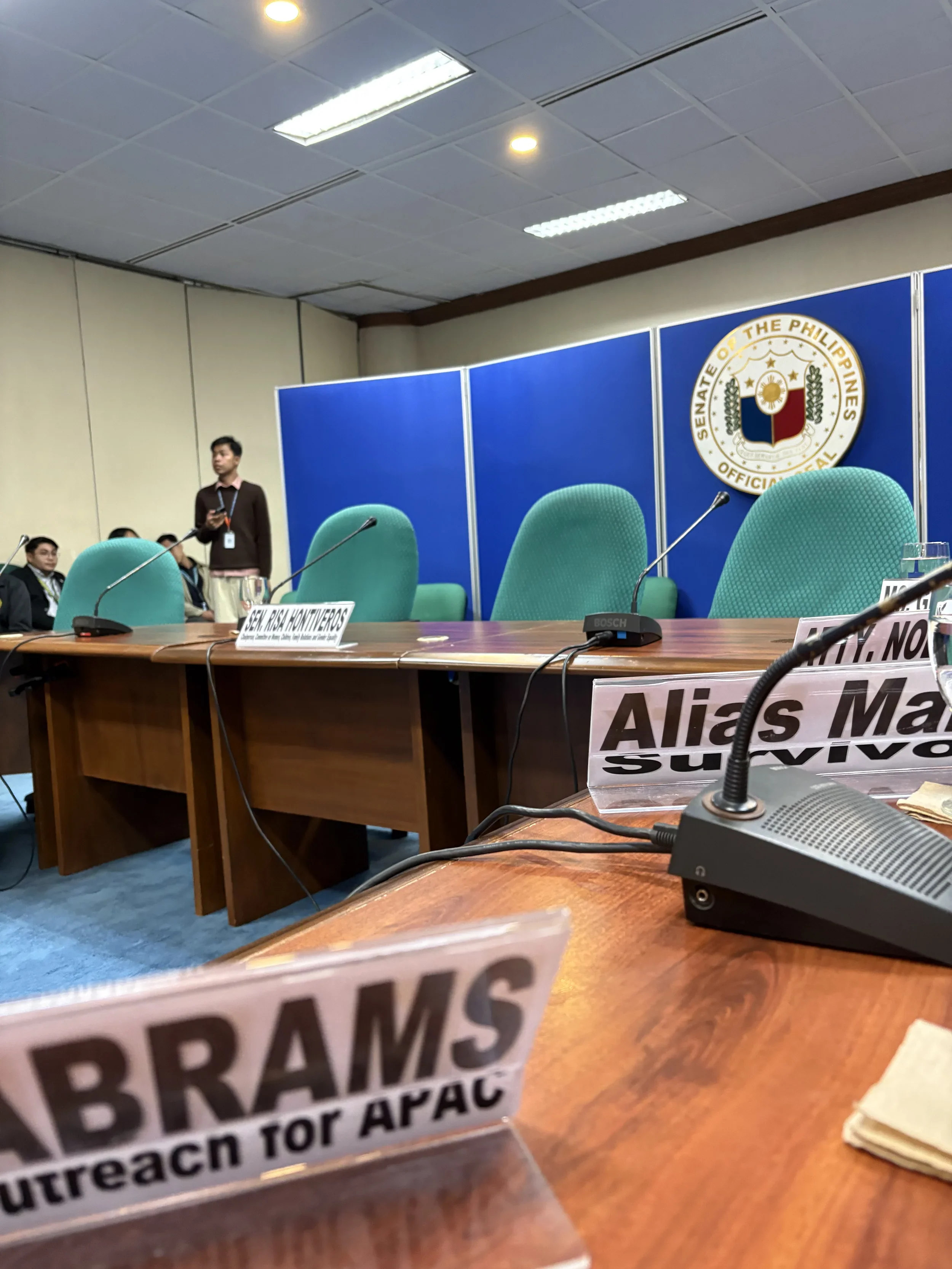 A conference room with a blue backdrop featuring the Senate of the Philippines seal. There are green chairs and a wooden table with nameplates, microphones, and a conference speaker. Some people are present, and one person is standing near the back.