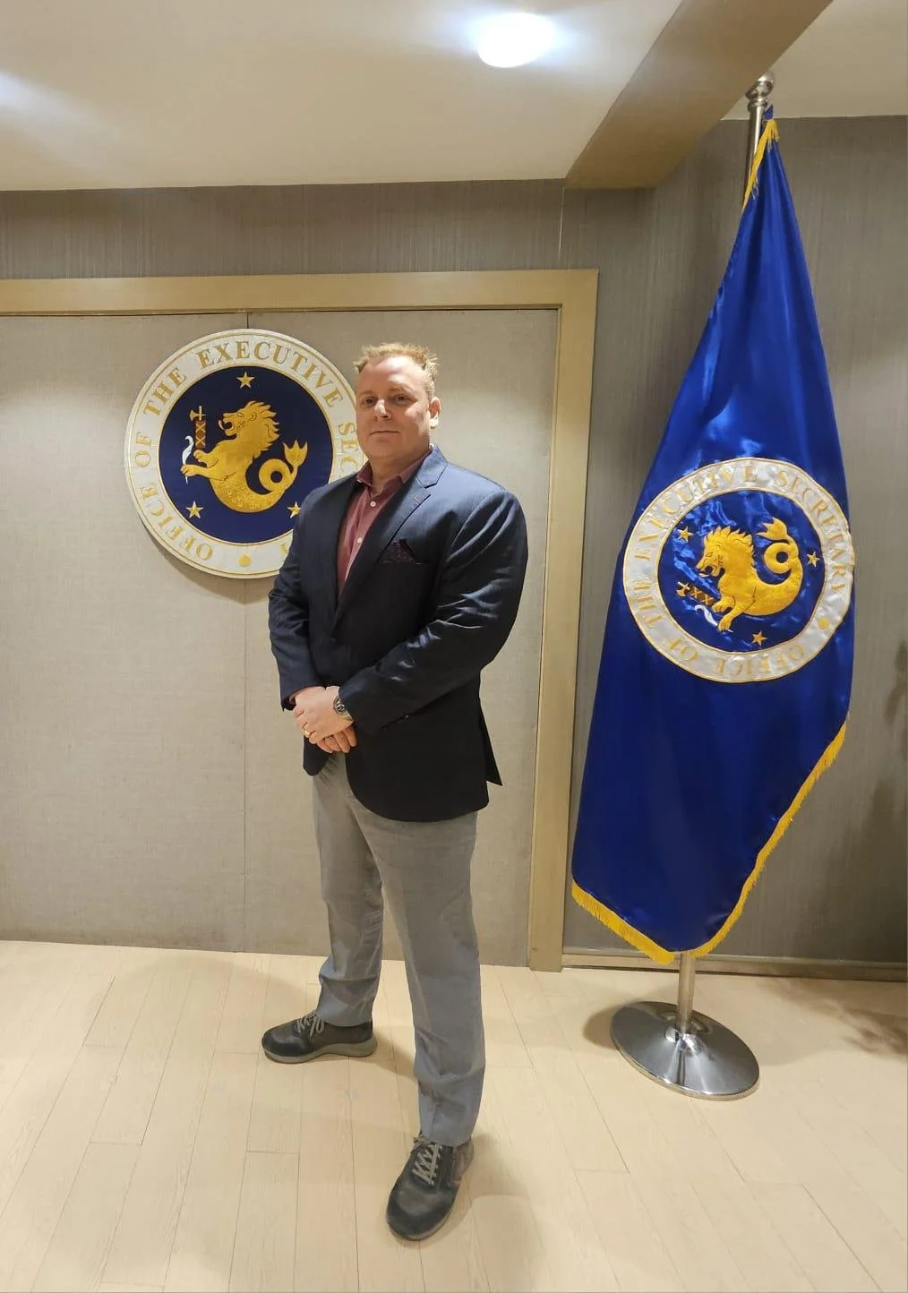 Man in business attire standing in front of a seal reading "Office of the Executive Secretary" and a blue flag with a circular emblem featuring a lion and stars.