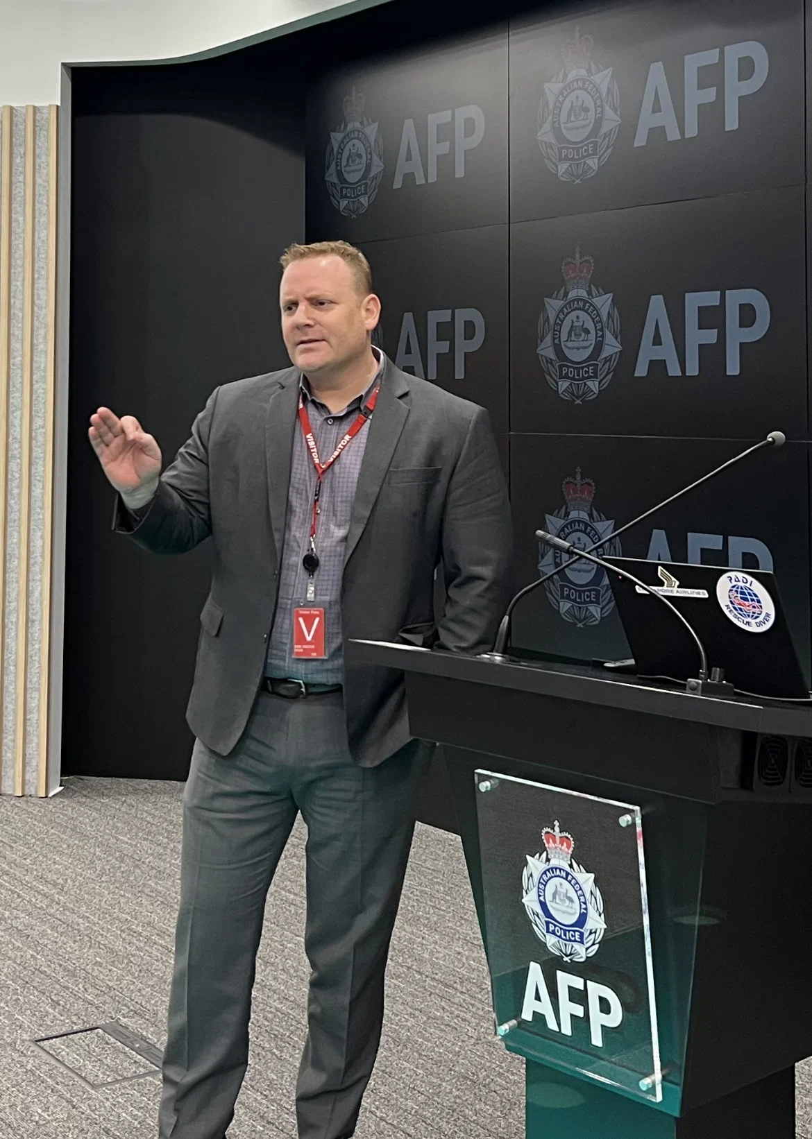 A man in a gray suit and dress shirt standing in front of a black background with the AFP and Australian Federal Police crest logo, speaking at a podium with AFP branding, during a media briefing or news conference.