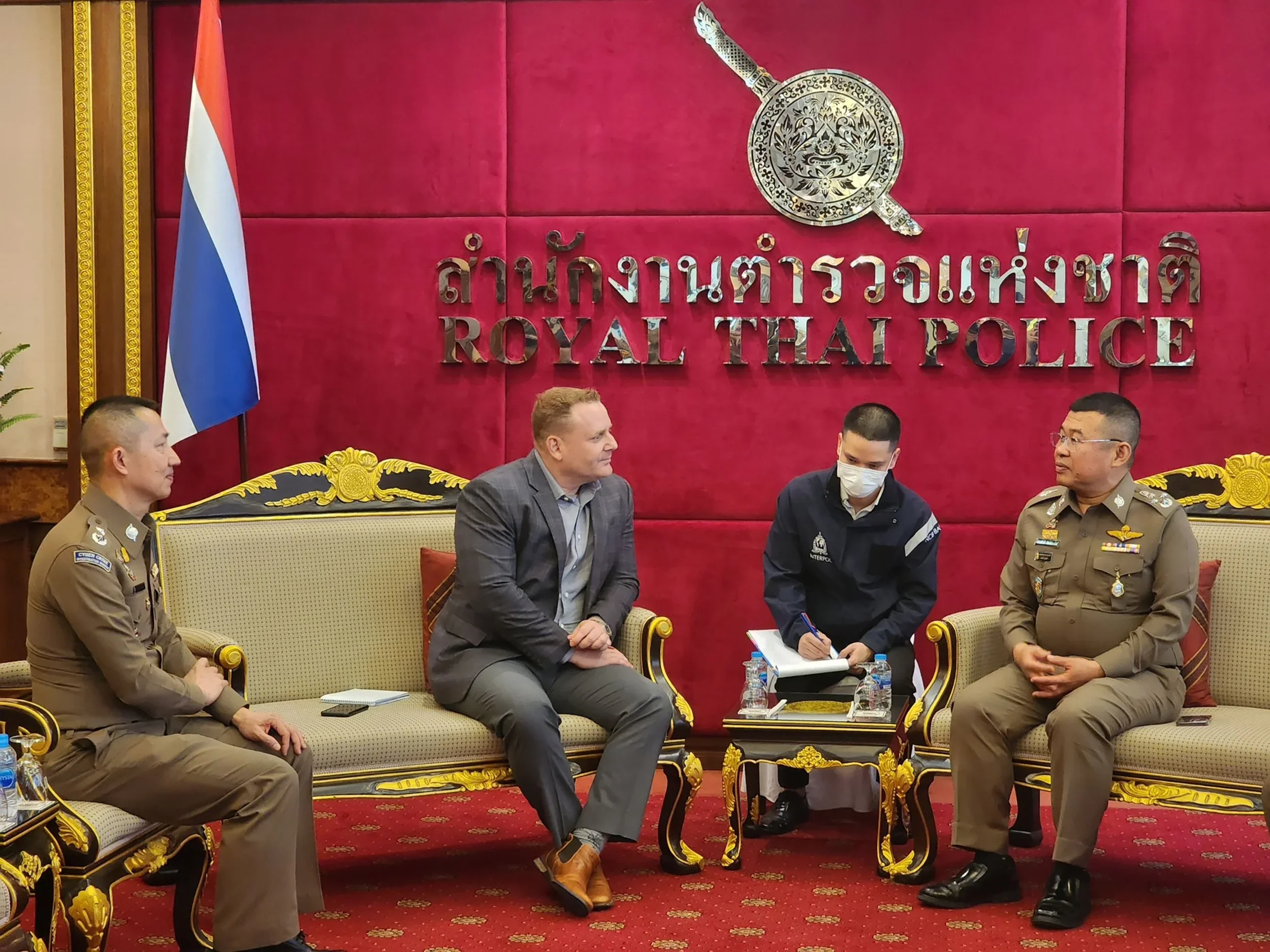 Four men in a meeting room with a red background and gold accents, including a sign that says 'Royal Thai Police' in English and Thai, with an emblem above.