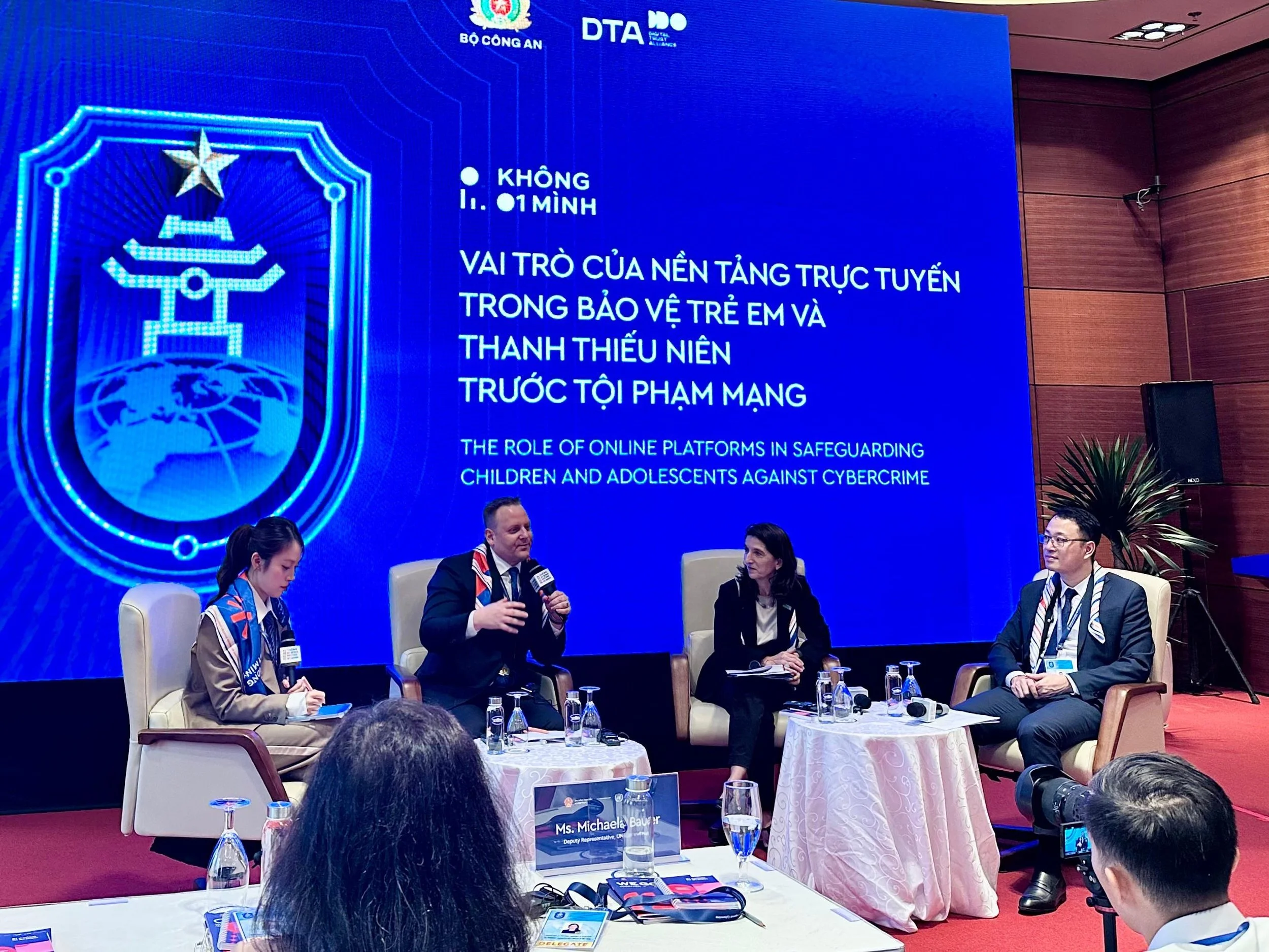 A panel discussion with four individuals seated on stage in front of a large blue screen displaying text in Vietnamese and English about online platforms' role in protecting children and adolescents against cybercrime. The panel includes three men in