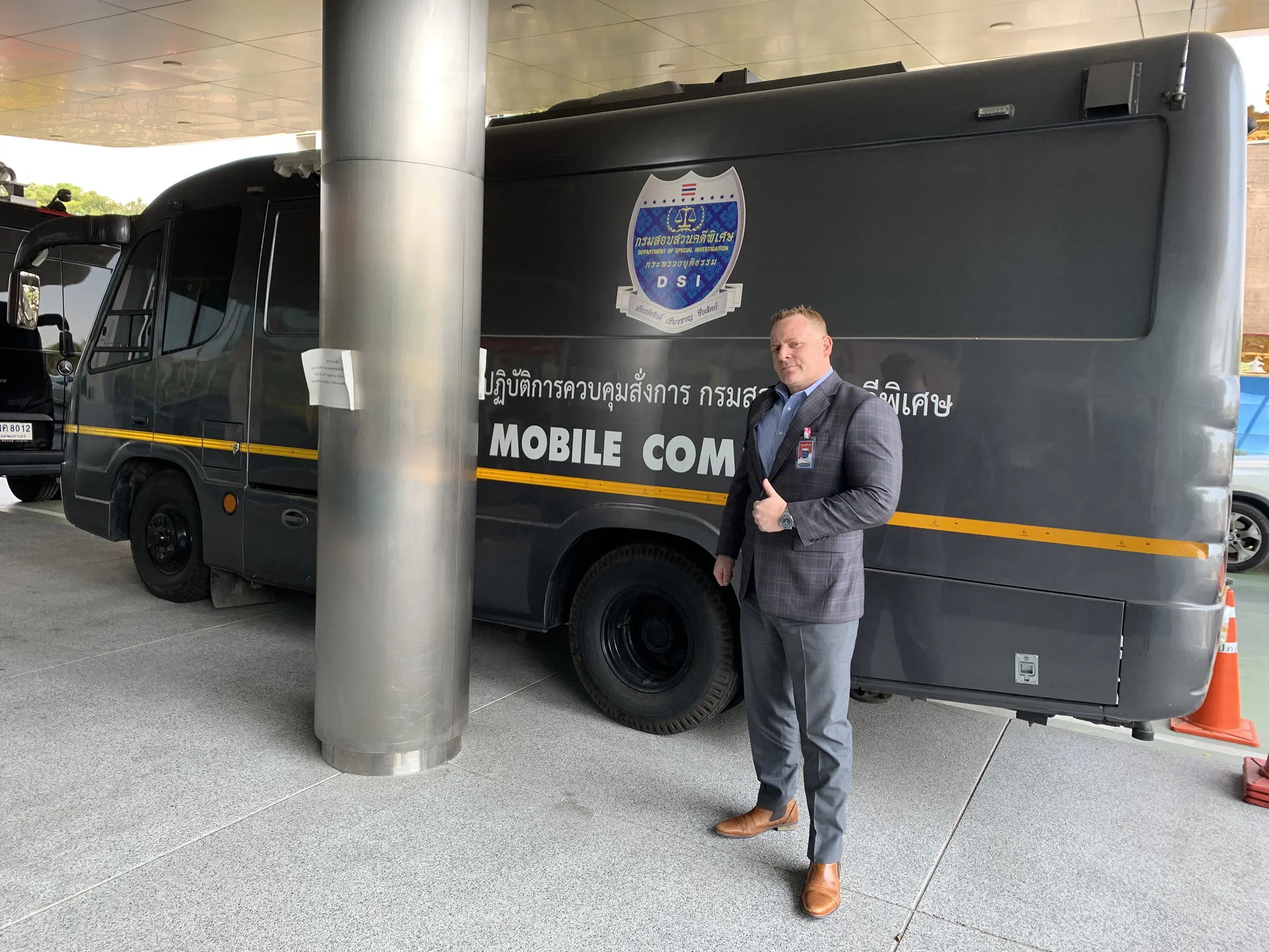 A man in a gray plaid suit standing in front of a large mobile command vehicle with a security badge on the chest, giving a thumbs-up gesture. The vehicle is black with official markings and text in a non-English language, and a logo on the door.