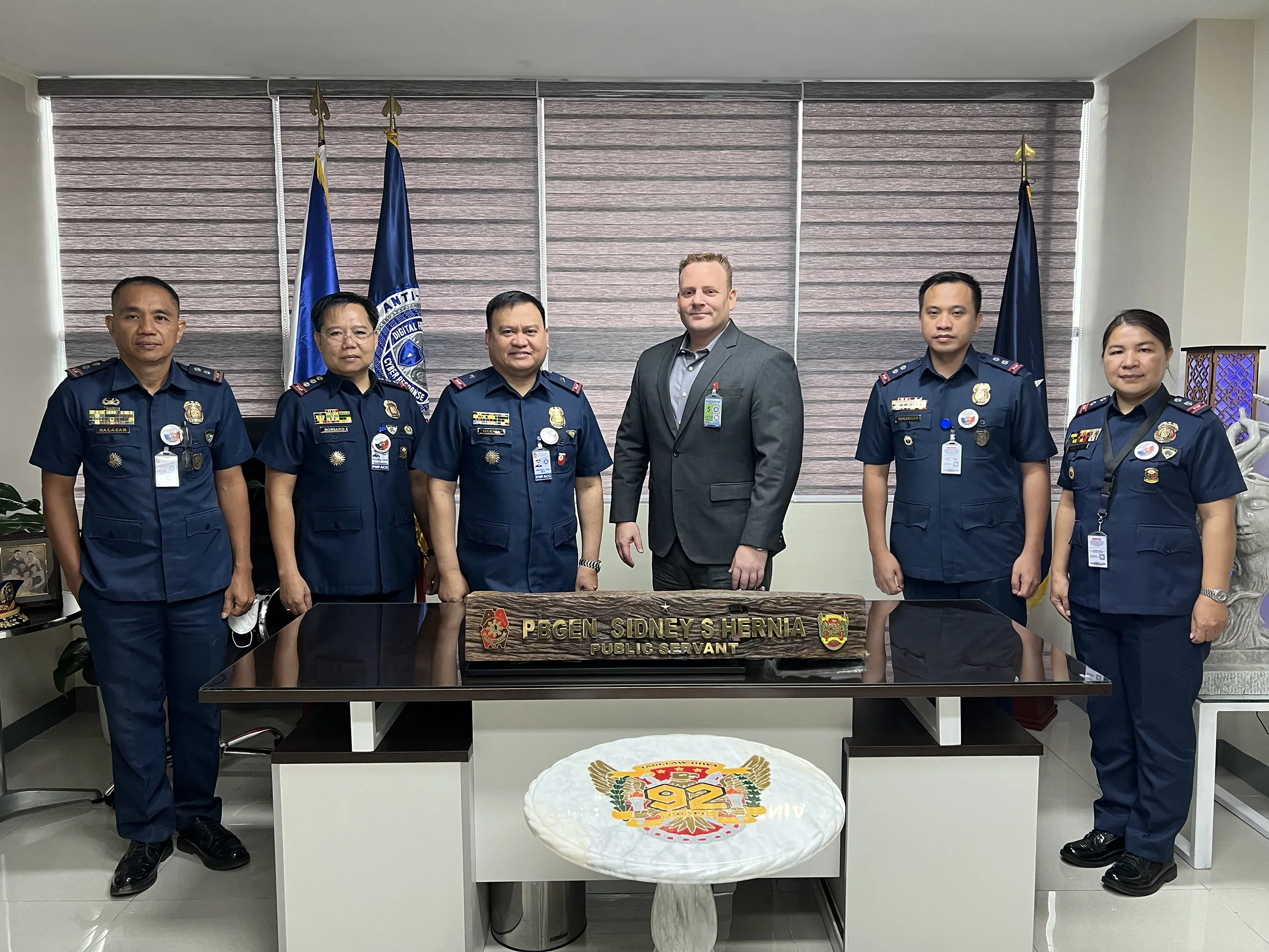 Group of six uniformed police officers and one man in a suit standing behind a desk in an office, with flags and a window with blinds in the background.