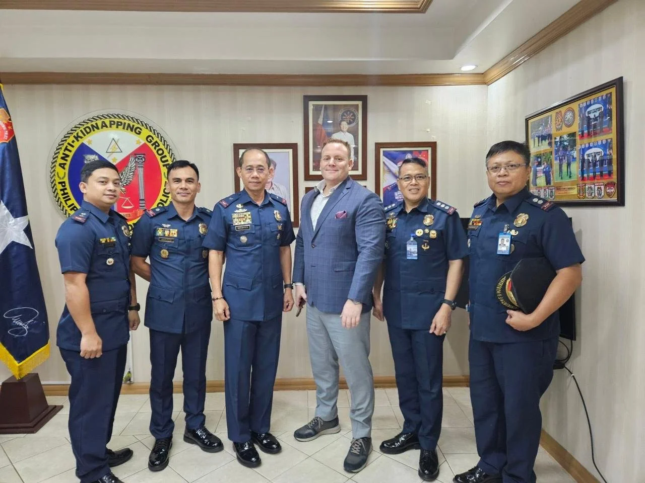 Six men standing together in a room, four wearing police uniforms and two in business attire, posing for a photo in front of a wall with framed pictures and a large police emblem.