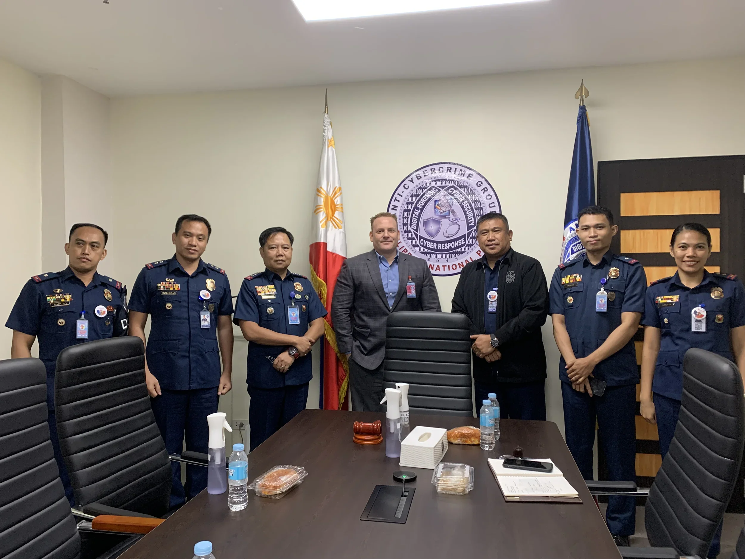 A group of seven uniformed personnel and two civilians standing in a conference room, posing for a group photo. Behind them are flags and a circular emblem on the wall that reads 'Anti-Cybercrime Group' and 'Cyber Response National'.