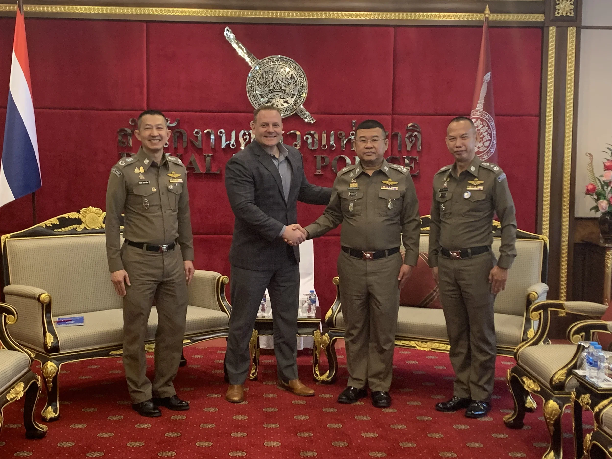 Four men, three in police uniforms and one in a business suit, shake hands in a formal setting with red walls, gold accents, and Thai flags in the background.