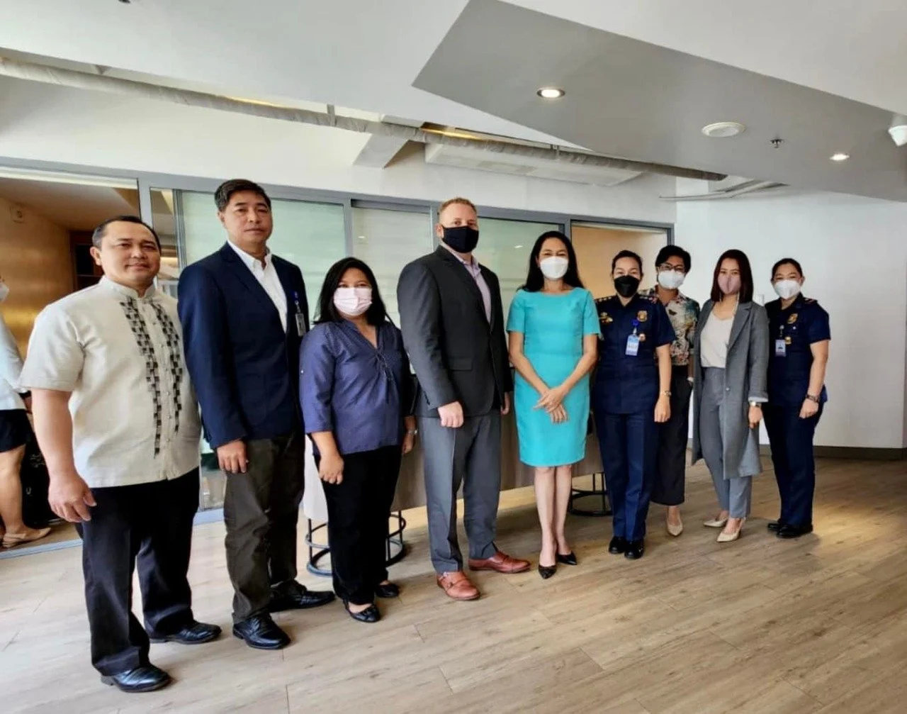 A group of nine people standing indoors, dressed in business and military attire, all wearing face masks.