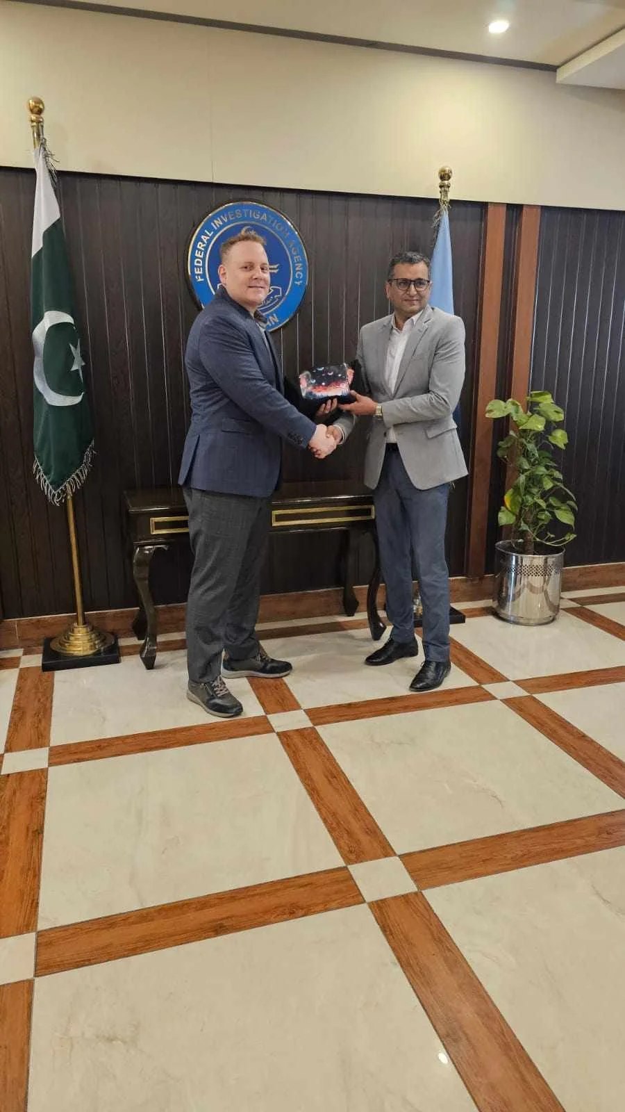 Two men in suits shaking hands and exchanging a gift in a formal setting with the federal investigation agency seal and Pakistani flag in the background.