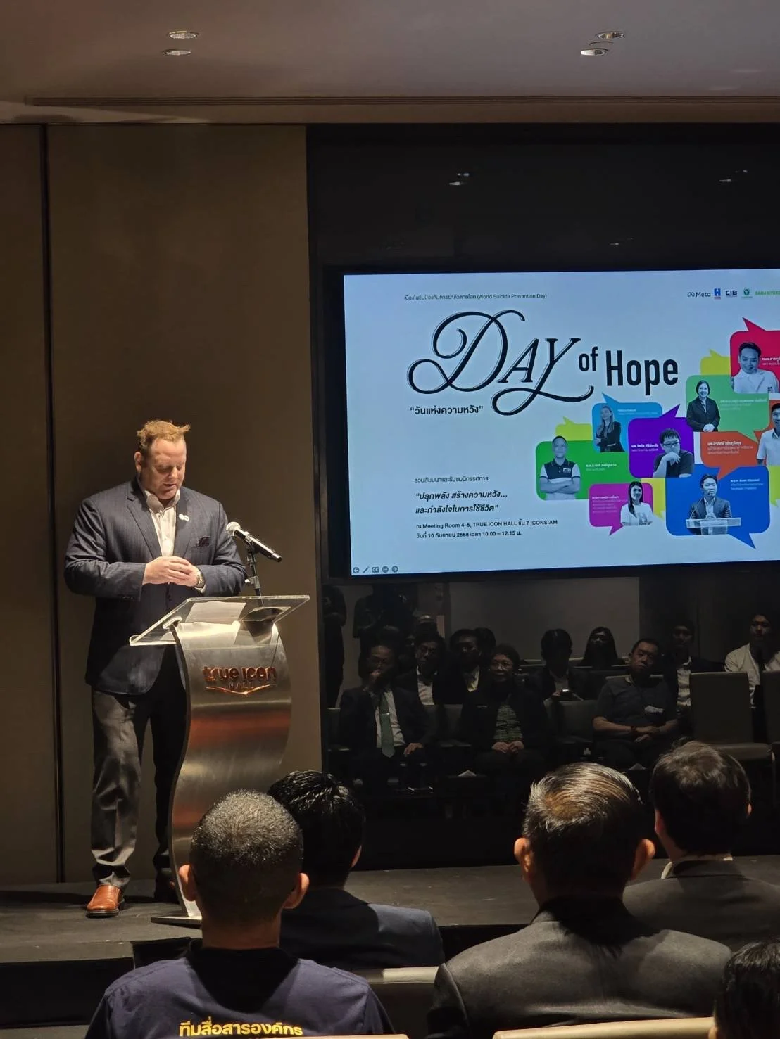 A man in a suit giving a speech at a podium during a conference or event, with an audience and a large screen displaying the event's title, "Day of Hope."