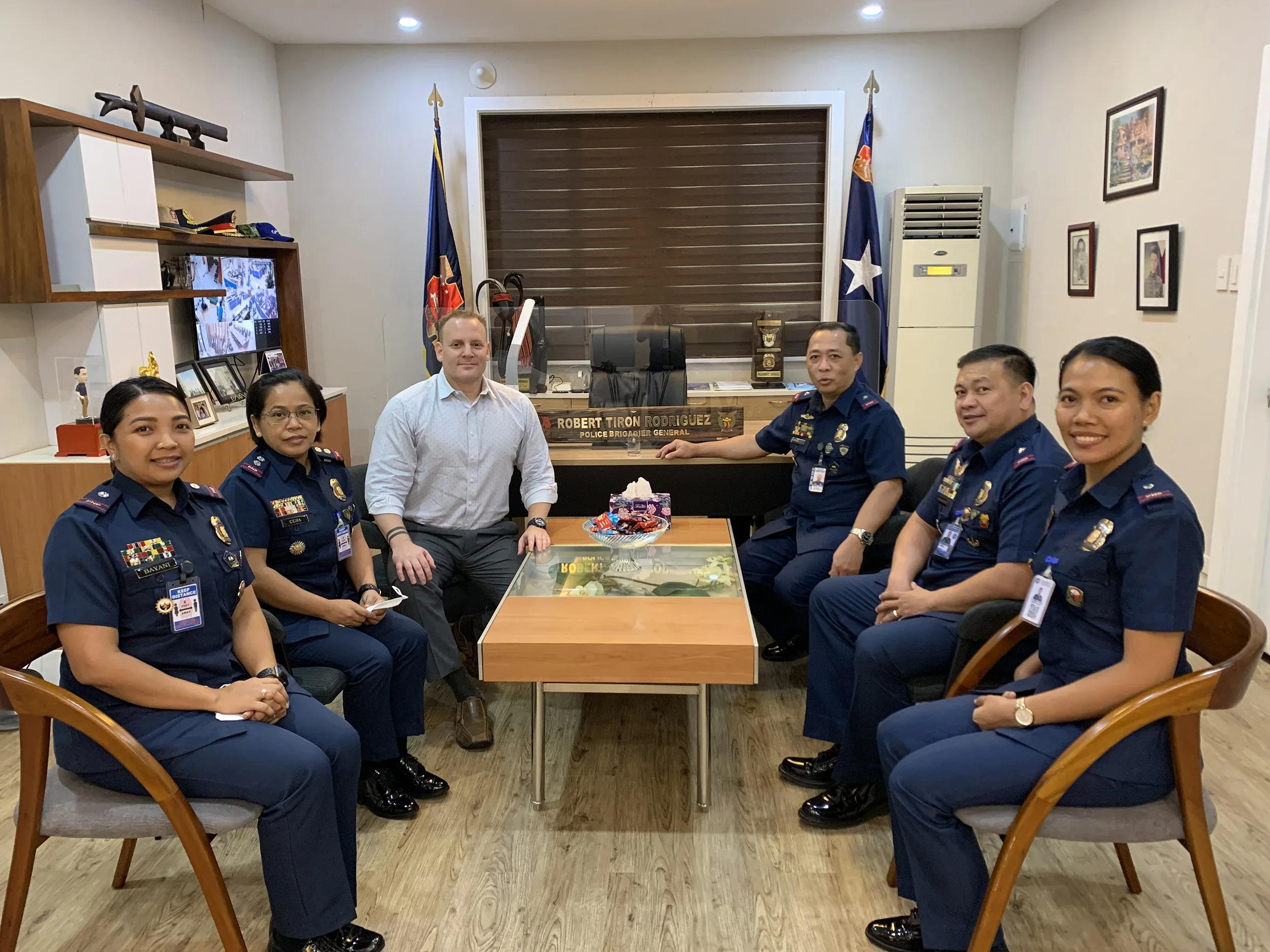 Six people, five in police uniforms and one in a business shirt, seated around a conference table in an office with Texas flags, framed pictures, and a wooden shelf with awards and medals.