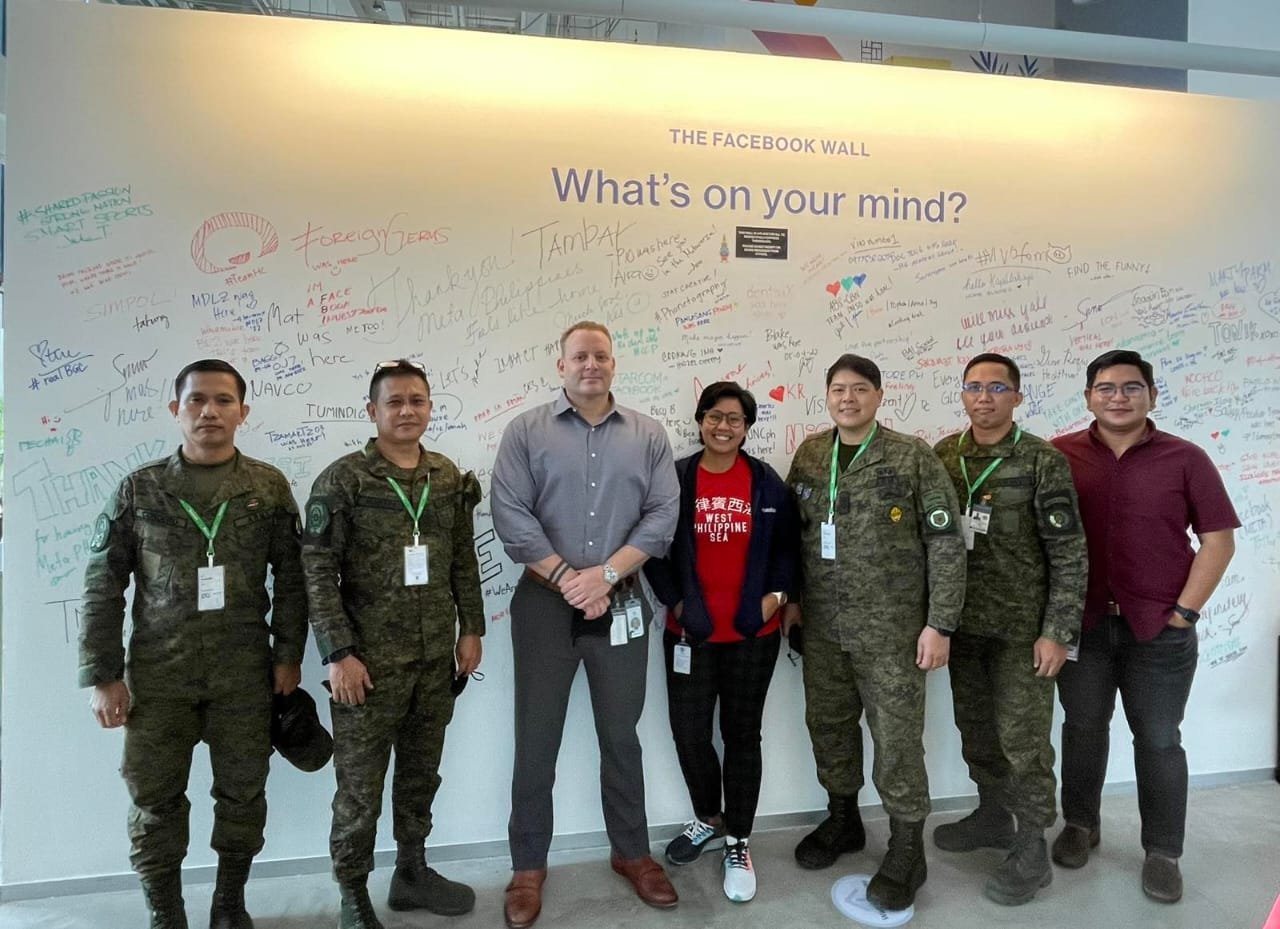 Group of seven people standing in front of a white wall covered with handwritten notes and signatures, with a large heading that reads 'What's on your mind?'