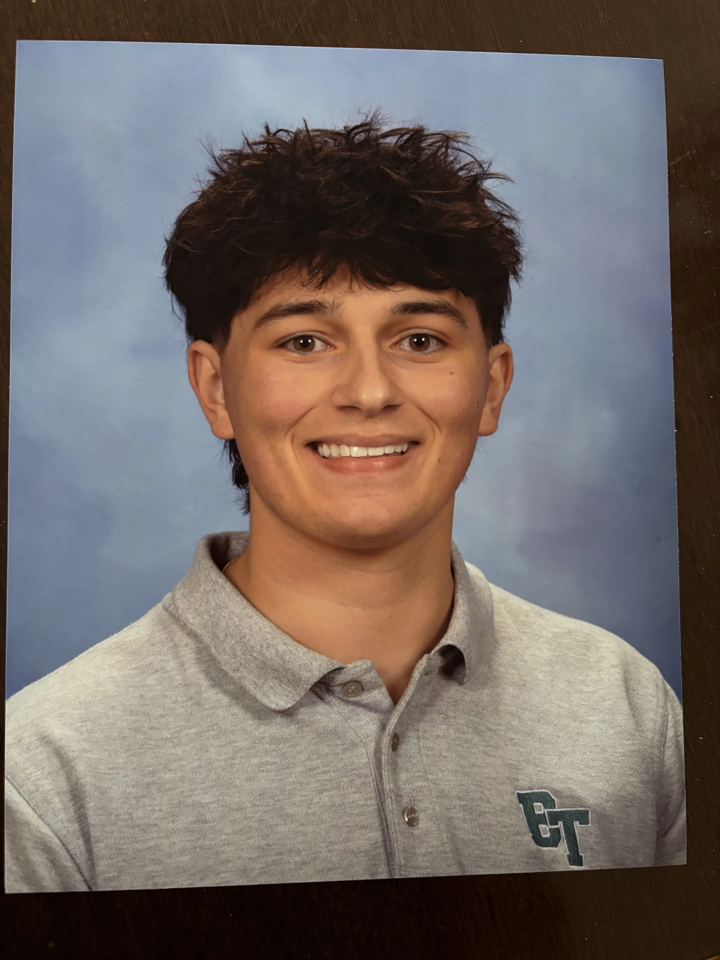 A young man smiling, wearing a light gray polo shirt with a 'GT' logo, against a blue background.