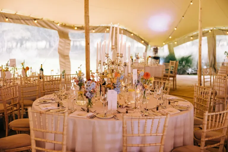 Wedding reception table with elegant floral centerpieces, candles, and neatly arranged place settings inside a decorated tent with string lights.