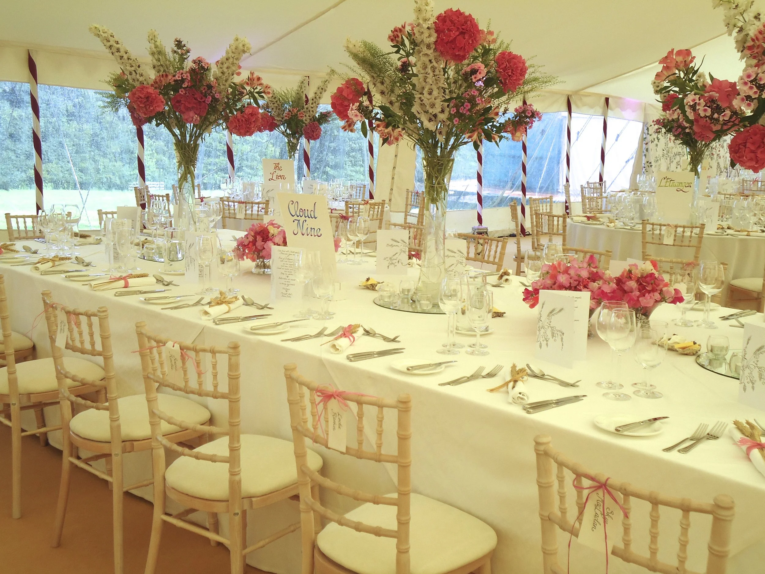Elegant dining setup with long table, beige chairs with white cushions, pink and white floral centerpieces, glassware, silverware, and white tablecloths inside a tent with large windows.