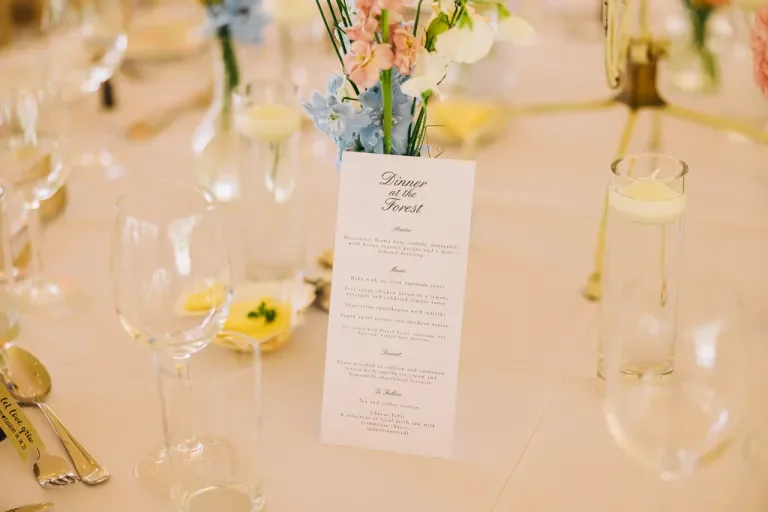 Elegant dining table setup with a floral centerpiece, a printed dinner menu, wine glasses, and candles.