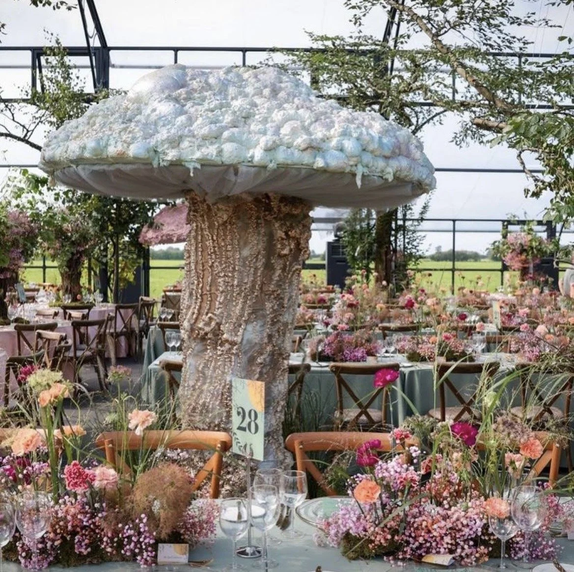 Indoor wedding or event venue decorated with pink and purple flowers, with a large mushroom-shaped centerpiece, and tables set with glassware and flowers.