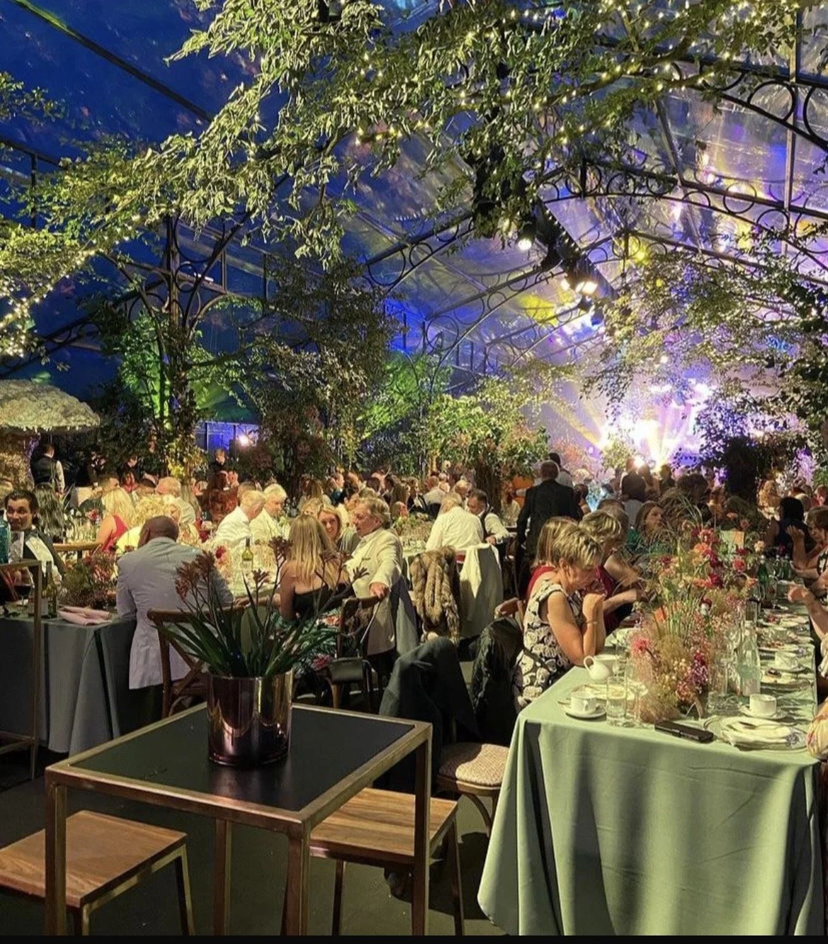 Indoor banquet hall decorated with greenery and colorful lights, filled with guests dining at tables with floral centerpieces.