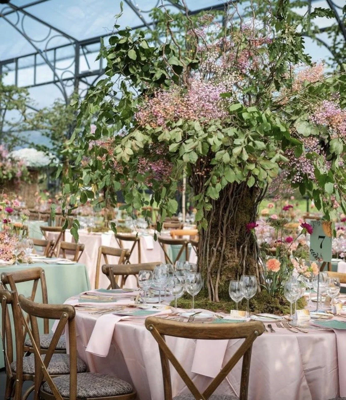 Elegant indoor event space decorated with a large floral centerpiece resembling a tree, table settings with glassware, cutlery, and pastel-colored tablecloths, surrounded by chairs and additional floral arrangements.