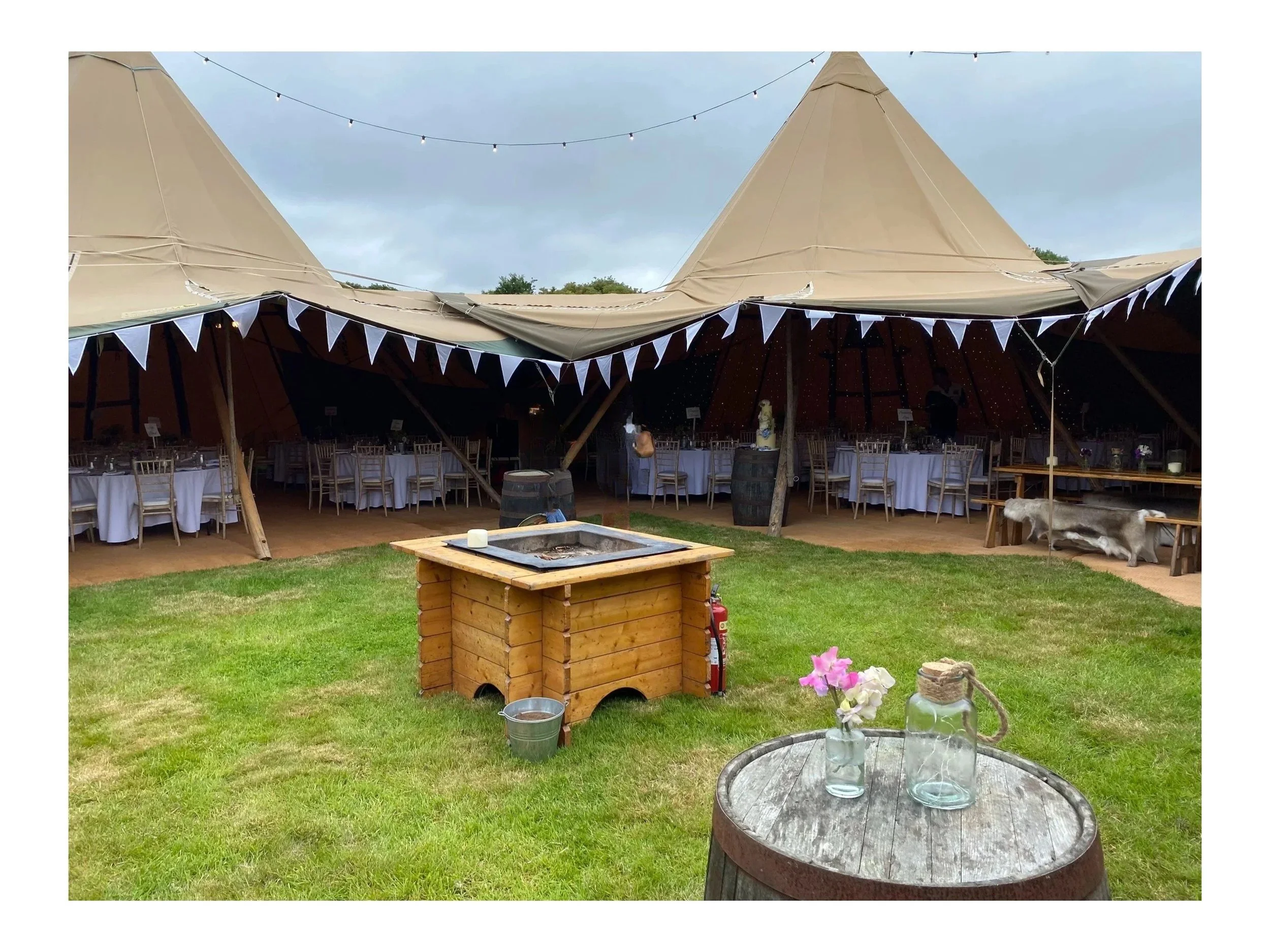 Outdoor event setup inside a large beige tent with white bunting and string lights, featuring tables with white tablecloths and beige chairs, and a grassy area with a wooden fire pit, a flower vase, and a barrel.