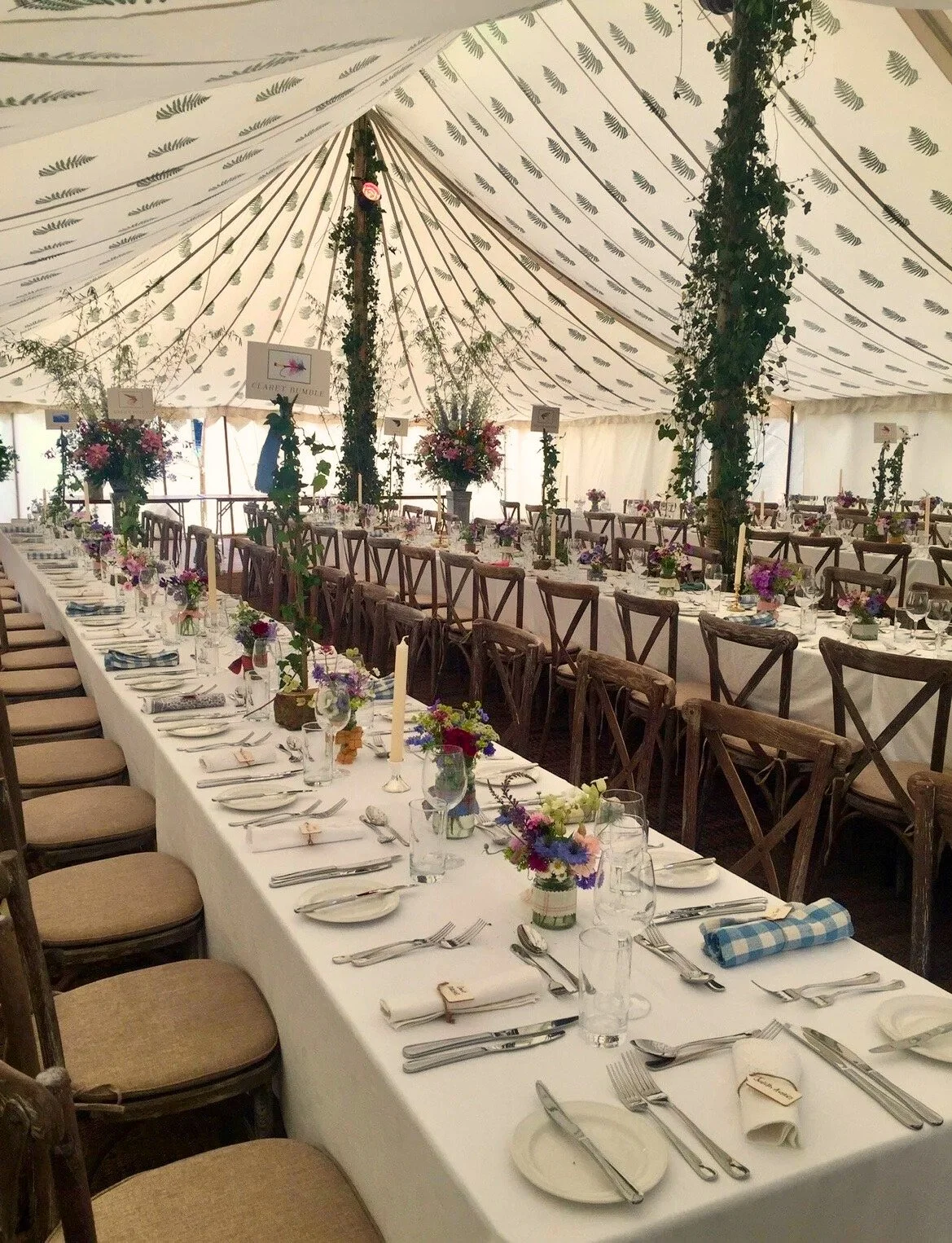 Long wedding reception table with white tablecloth, floral centerpieces, and place settings under a decorated tent with hanging greenery.
