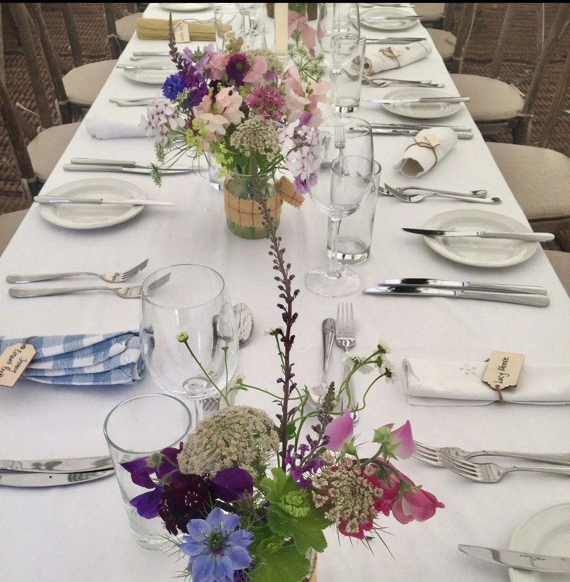 A decorated dining table with a white tablecloth, arranged with multiple sets of silverware, glassware, and porcelain plates, featuring a central floral arrangement with pink, purple, and white flowers.